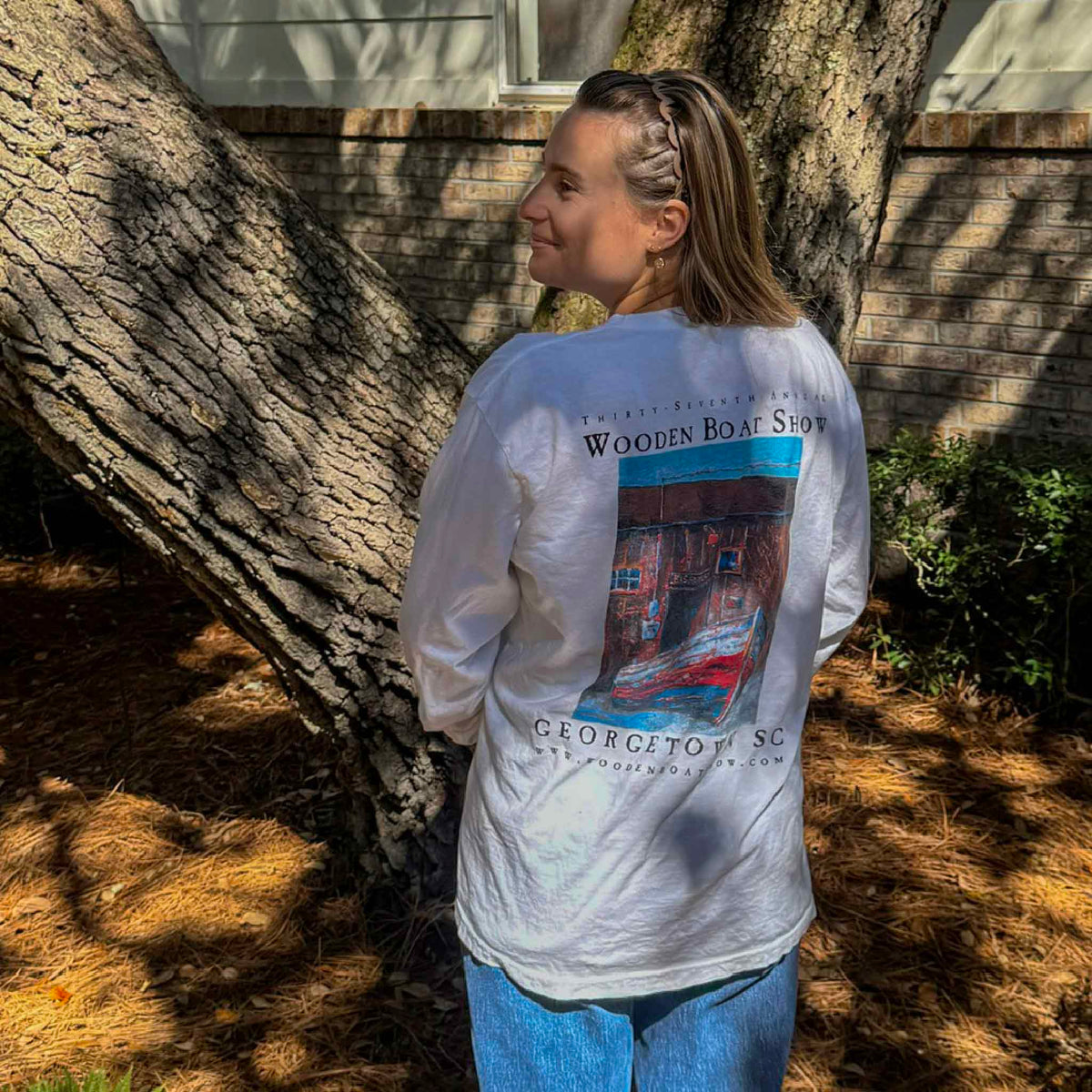 Kyra models the back of the official 2026 Georgetown Wooden Boat Show white Comfort Colors long sleeve T-shirt beneath a large oak tree at Gray Man Gallery in Pawleys Island, South Carolina. The back features local artist Vida Miller's colorful Fish House painting of a historic Georgetown waterfront building and wooden boat, commemorating the 37th Annual Wooden Boat Show.