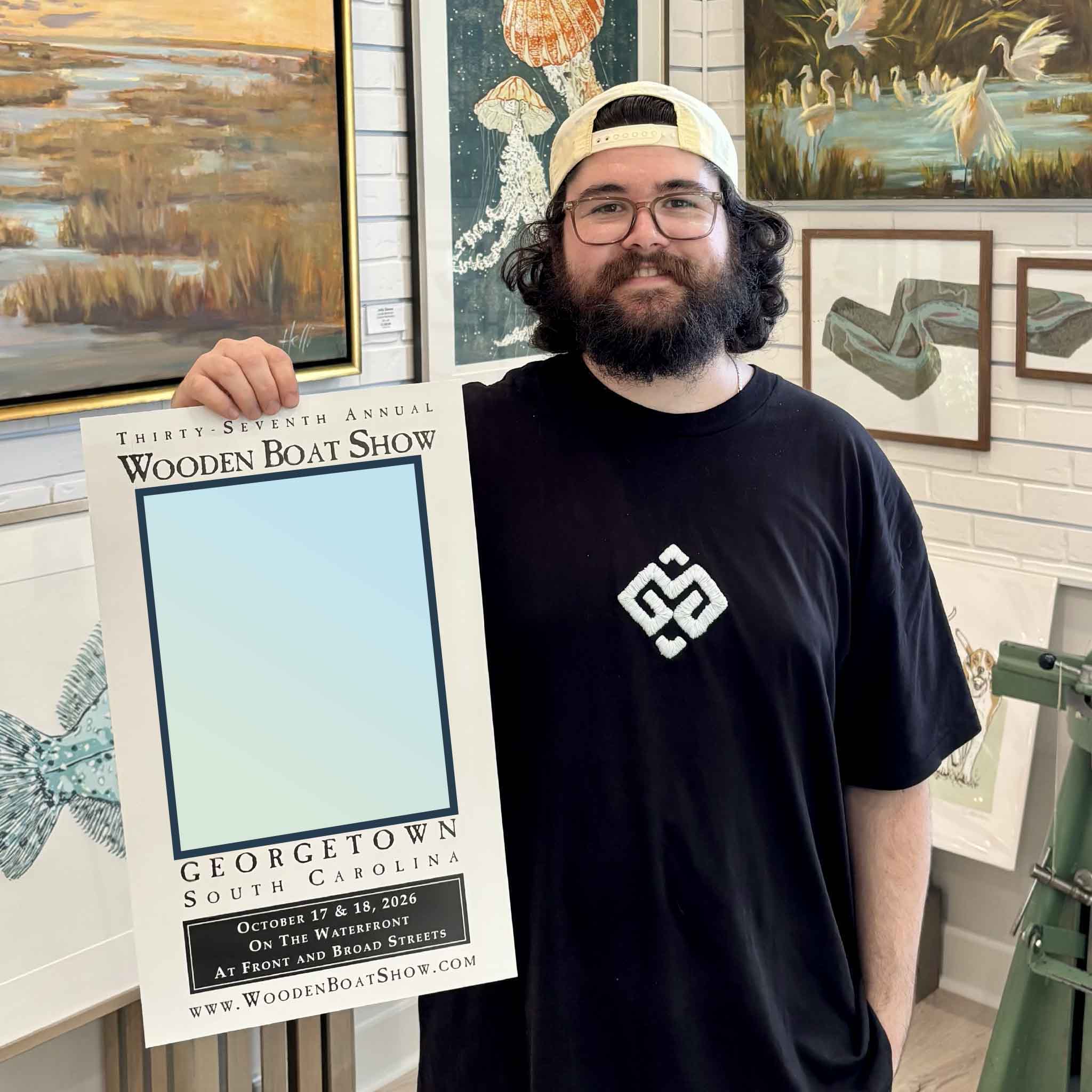 Peyton holds a teaser of the official 2026 Georgetown Wooden Boat Show poster inside Gray Man Gallery in Pawleys Island, South Carolina, with the artwork intentionally hidden ahead of tonight’s official poster and featured artist reveal.