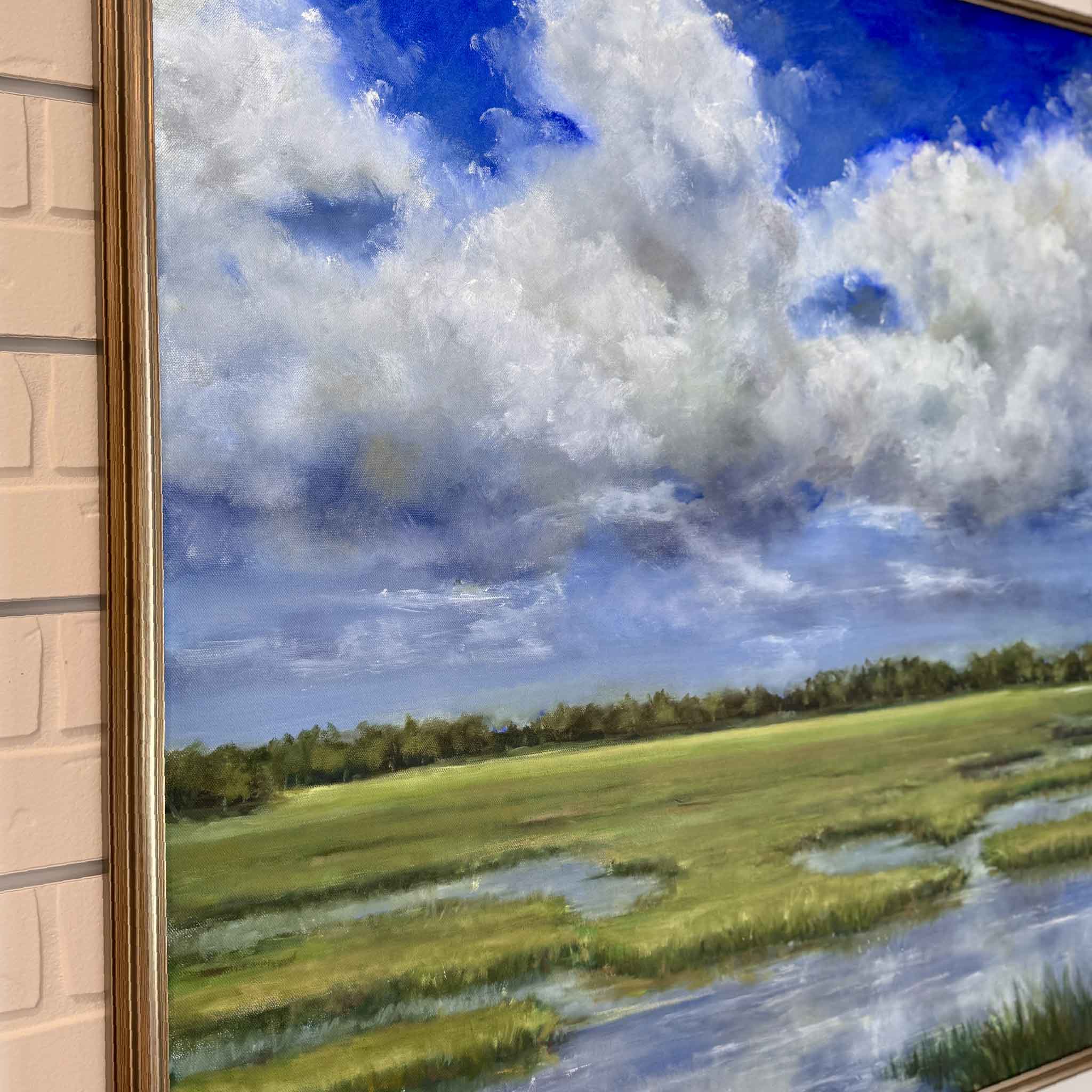 Framed oil painting of a serene Lowcountry marsh landscape under a dramatic sky filled with billowing white clouds. The scene features lush green grasses, reflective tidal pools, and a distant tree line, all set against a vibrant blue sky.