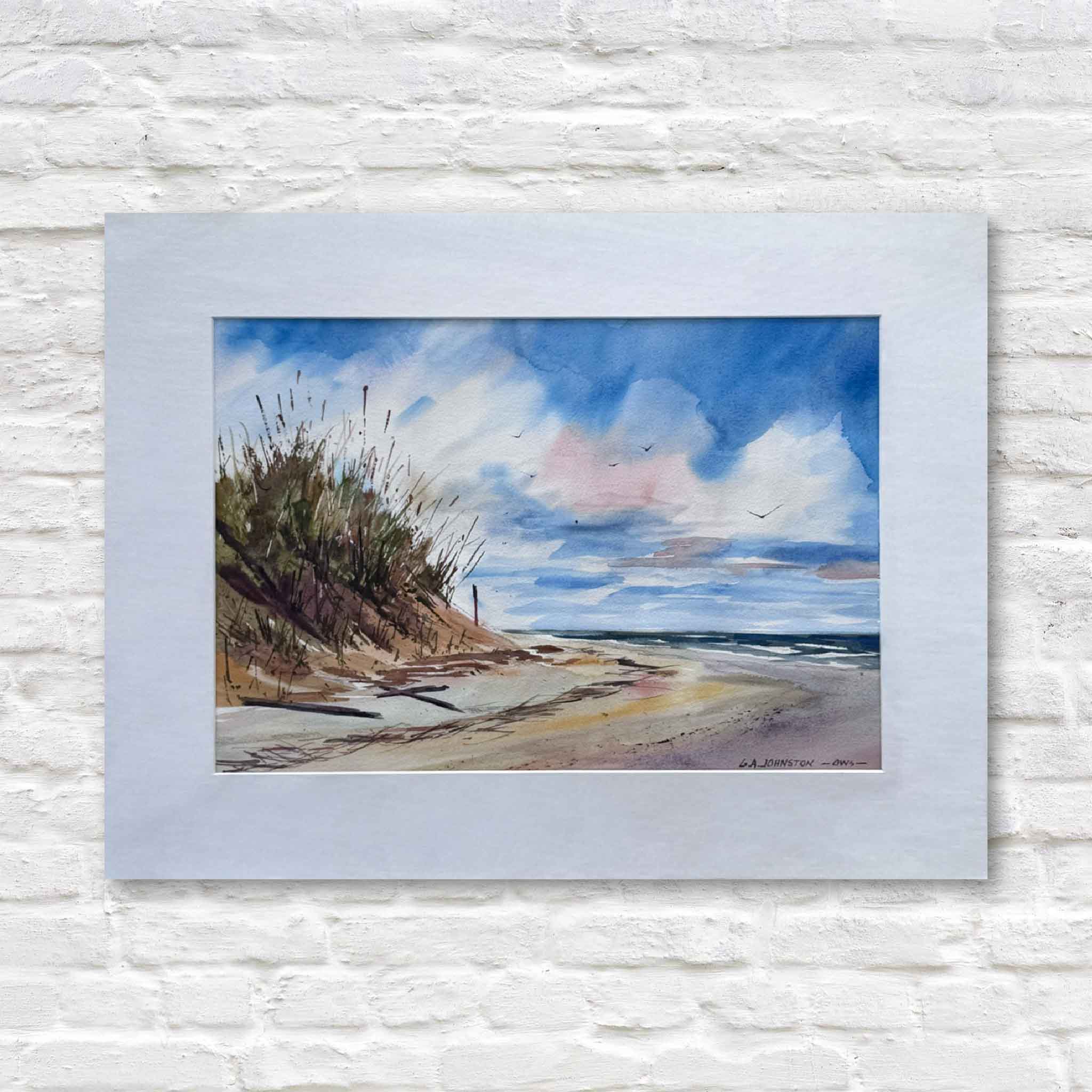 matted painting of a beach scene with a blue sky and clouds on a white brick wall.