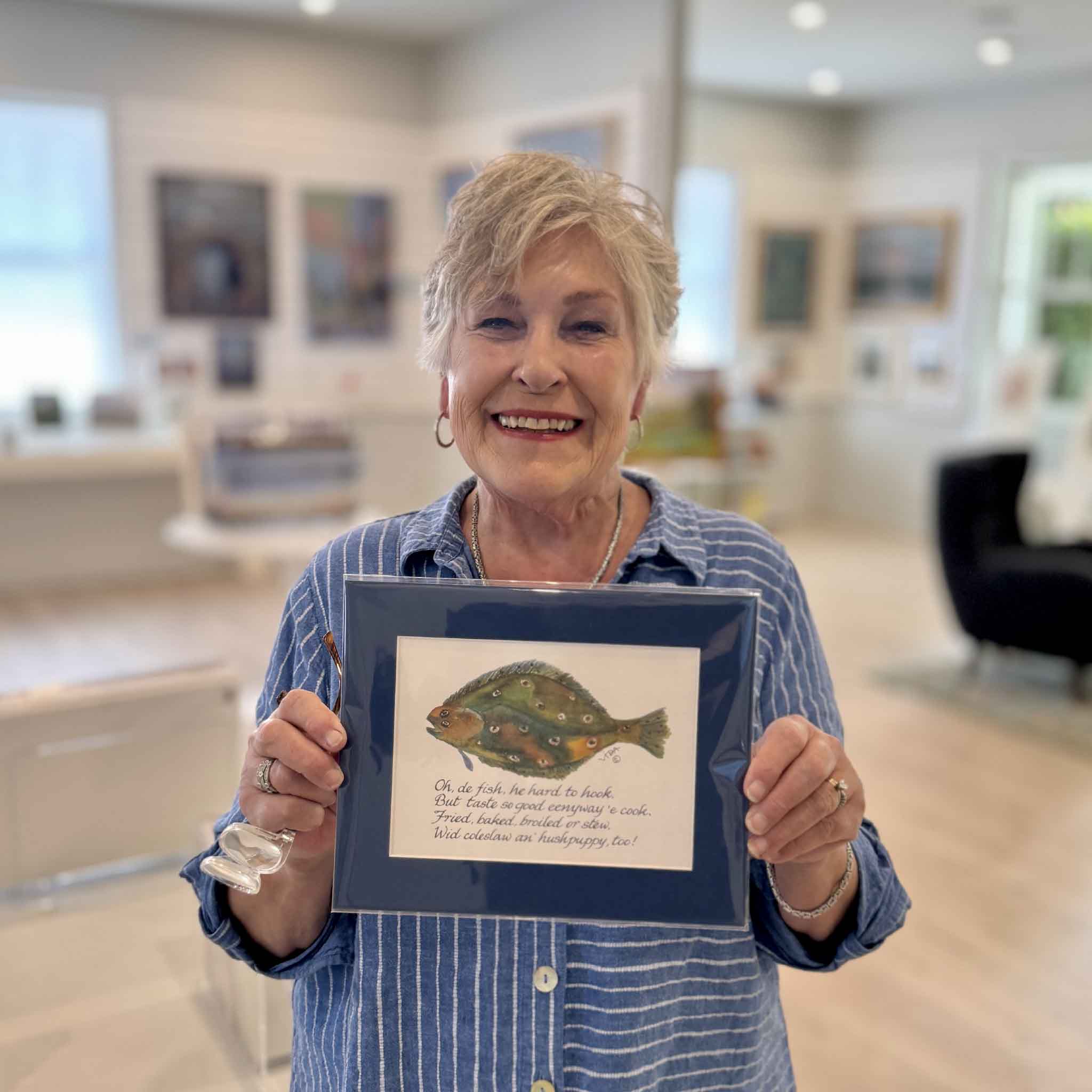 Local Pawleys Island artist, Vida Miller, smiling and holding a print of a fish illustration with a whimsical verse, on display at Gray Man Gallery.