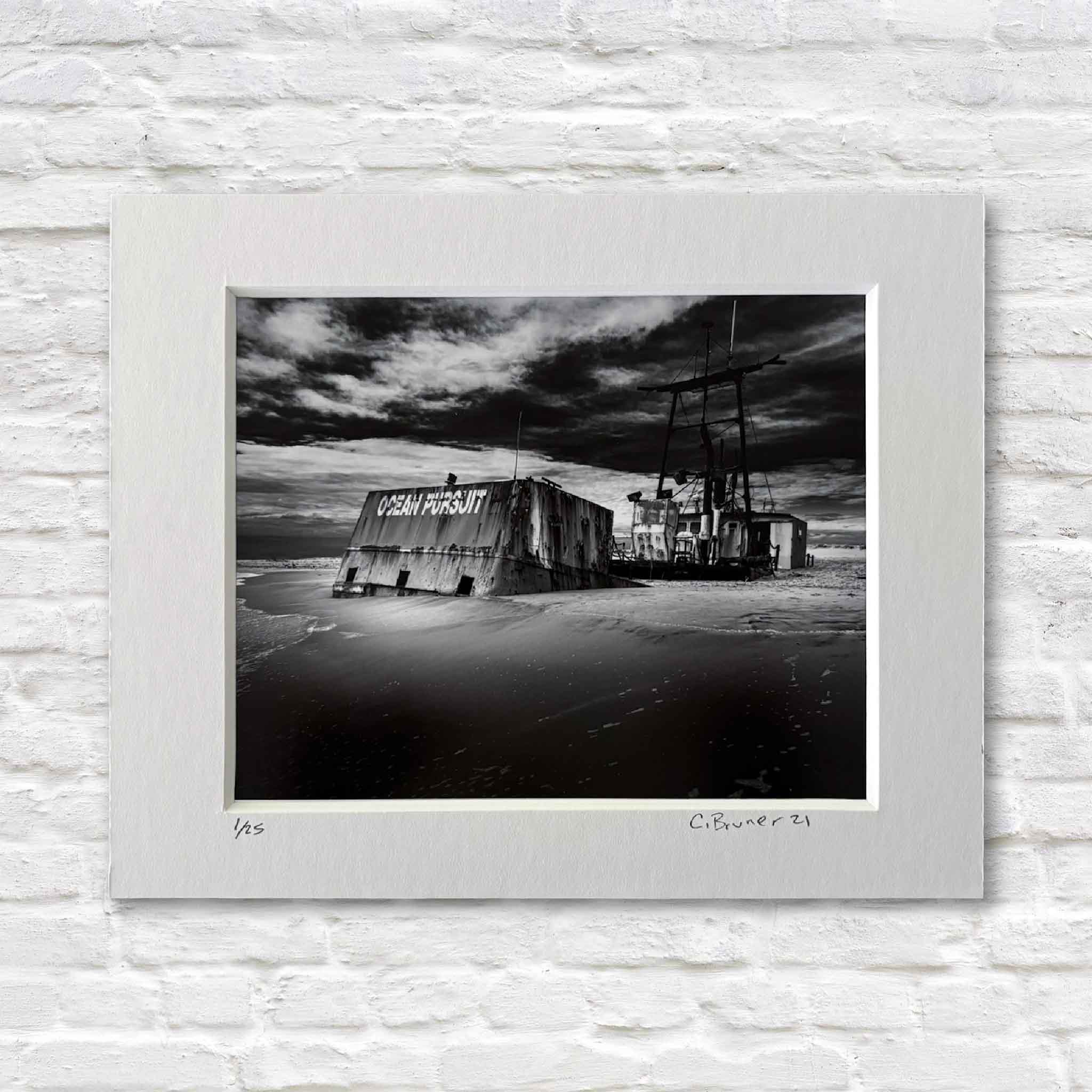 matted black and white photograph of a derelict shipwreck on a beach against a dramatic sky.