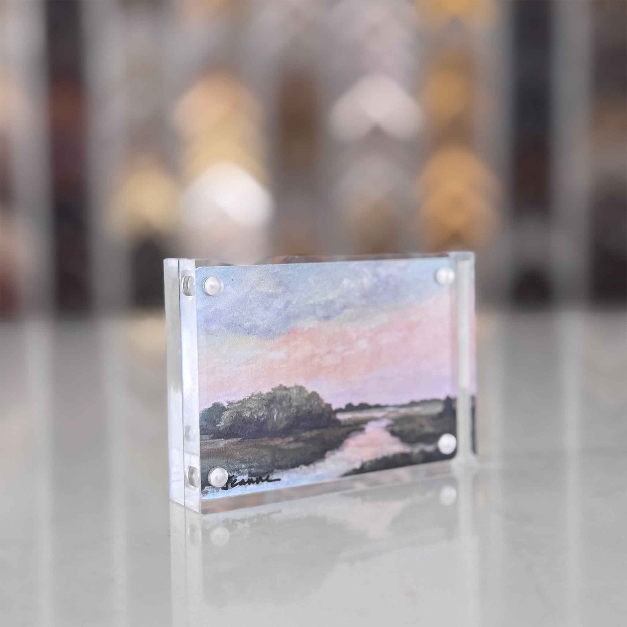 Small landscape painting in a clear frame on a reflective surface with a blurred background