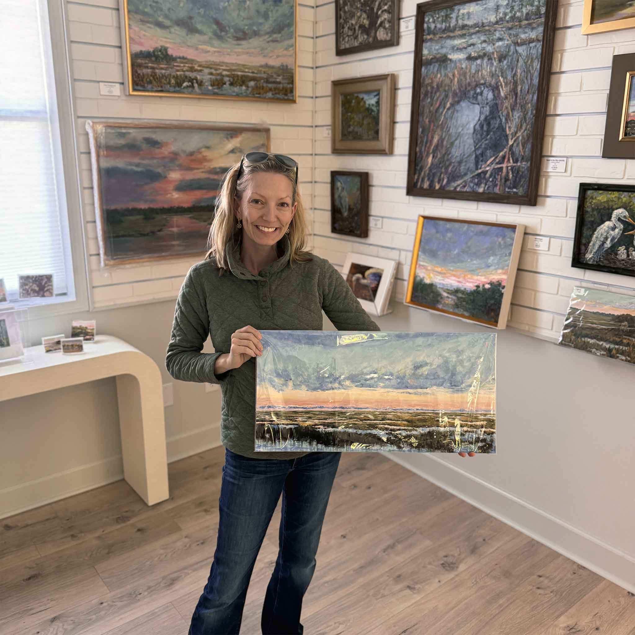 Jeanne O’Neal holding her coastal landscape print titled “Classy,” featuring a wide Lowcountry marsh view with dramatic sky colors.