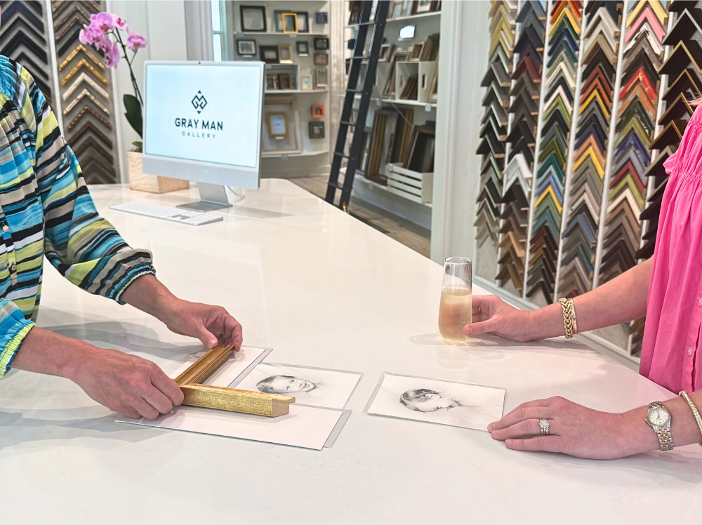 A framer presents a gold frame corner sample over a portrait while a client considers design options, surrounded by an array of frame samples at Gray Man Gallery.