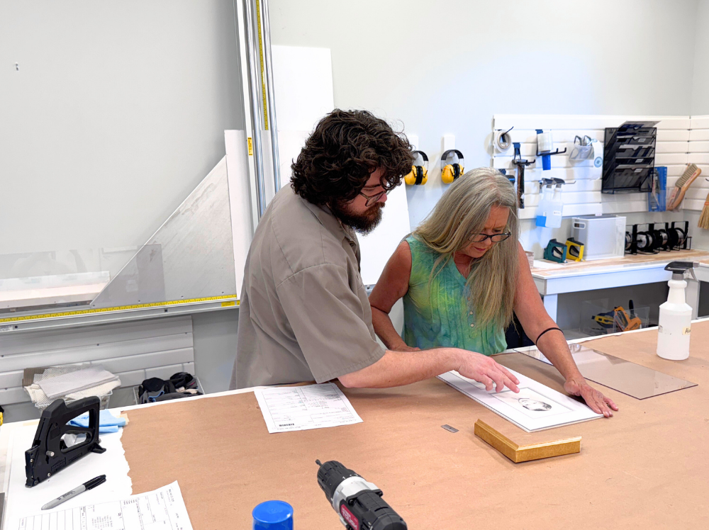 Two framers at Gray Man Gallery review and prepare artwork for mounting, with a mat and gold frame sample nearby, ensuring precise placement on the backing board.