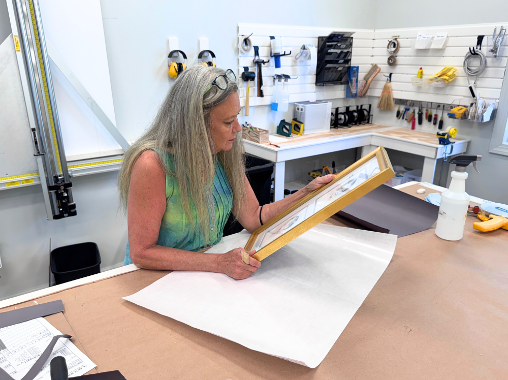 A framer at Gray Man Gallery closely inspects a completed, gold-framed artwork on the worktable, ensuring perfect alignment and quality before final packaging.