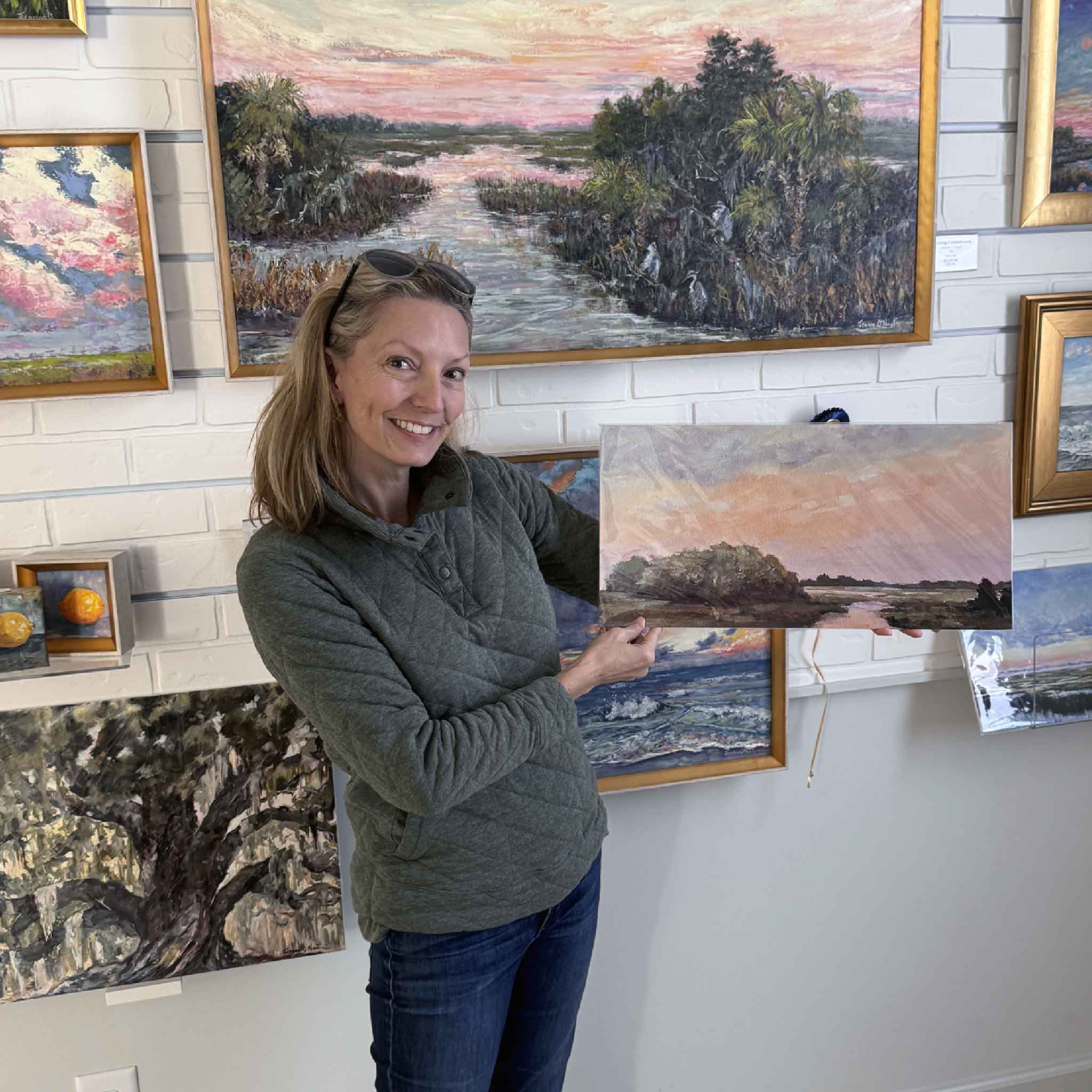 Local artist Jeanne O'Neal inside an art gallery holding a rectangular coastal landscape print featuring a soft pastel sunset and marsh scene. Several framed paintings hang on the wall behind her.