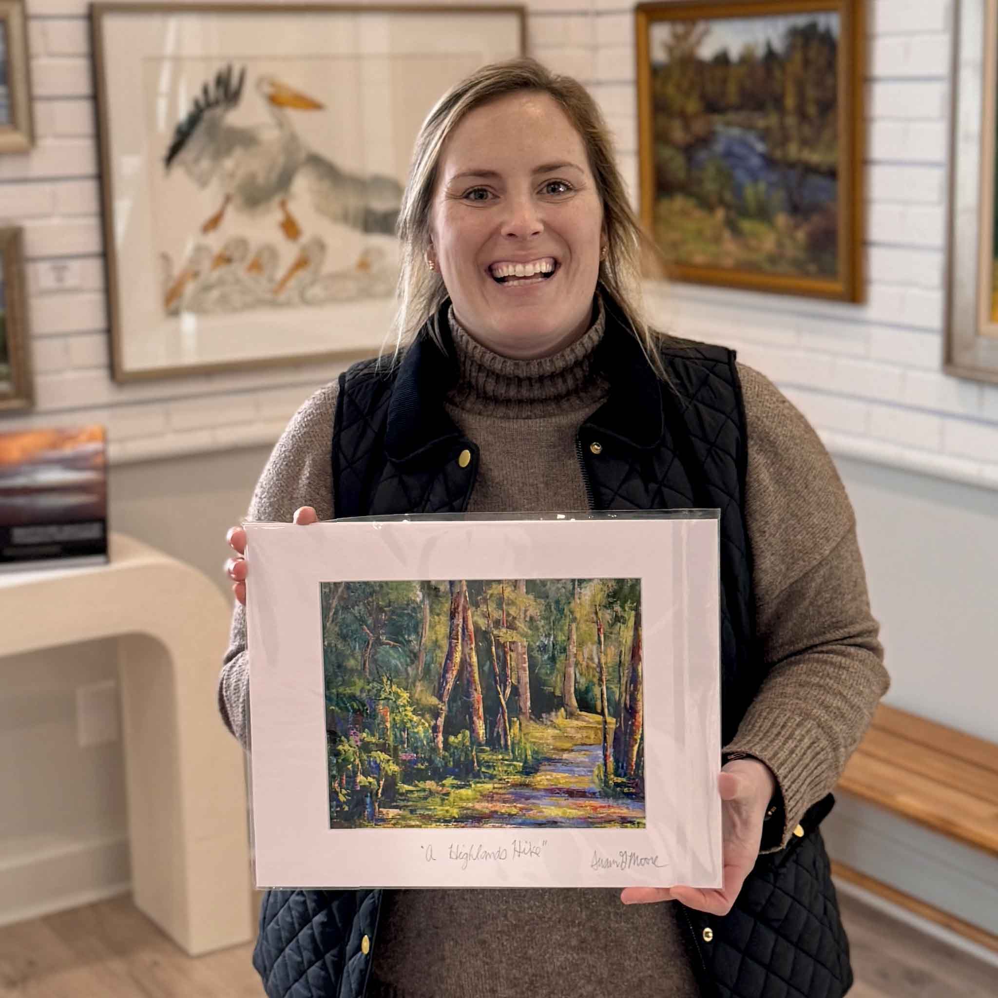 Bordy holding “A Highlands Hike” fine art giclée print by local artist Susan Moore inside Gray Man Gallery in Pawleys Island, showcasing matted 11x14 woodland trail artwork