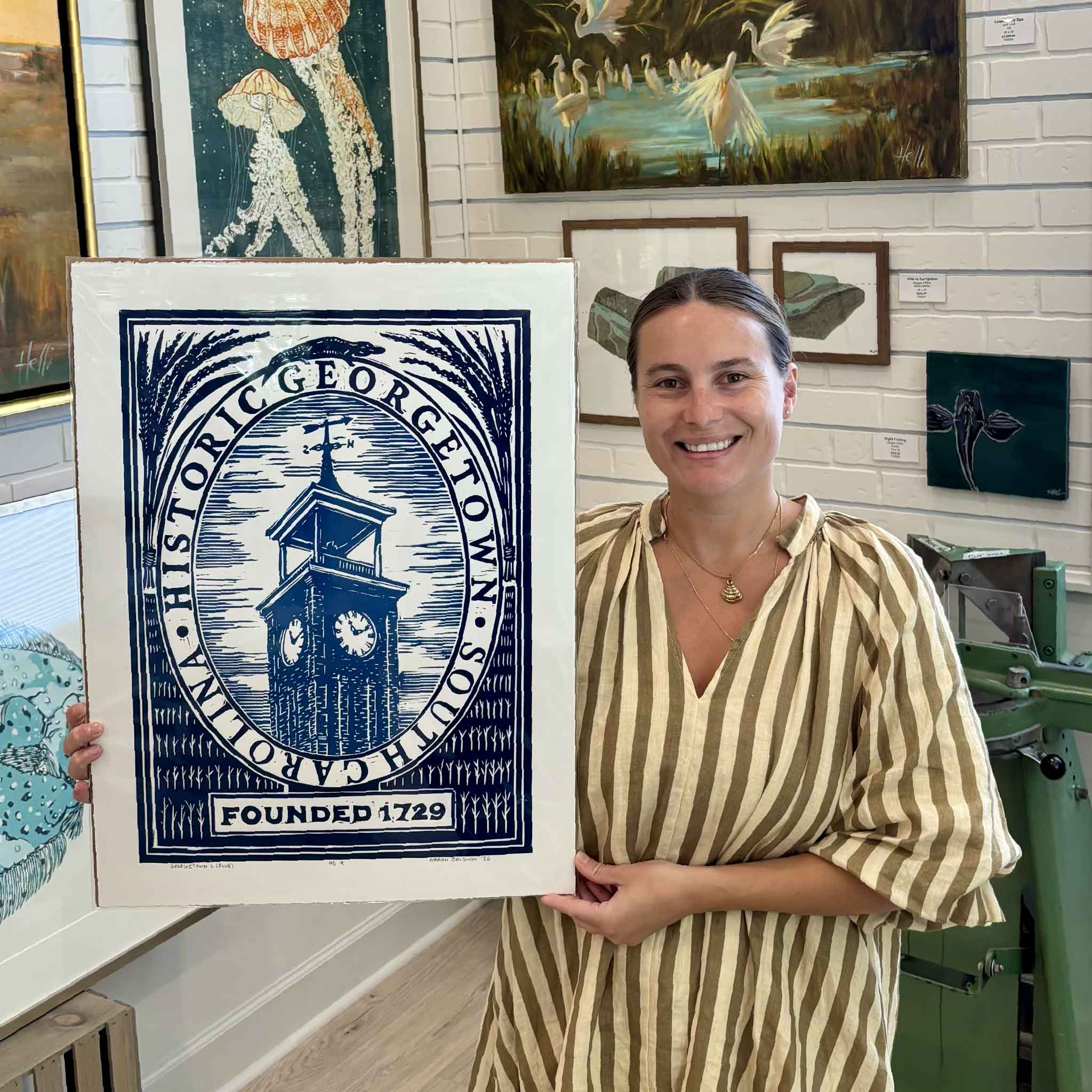 Kyra holding Georgetown by South Carolina artist Aaron Baldwin, an original hand-pulled blue linocut depicting the historic clock tower in downtown Georgetown, South Carolina, with an alligator detail above the tower and “Founded 1729” below, photographed inside Gray Man Gallery in Pawleys Island.