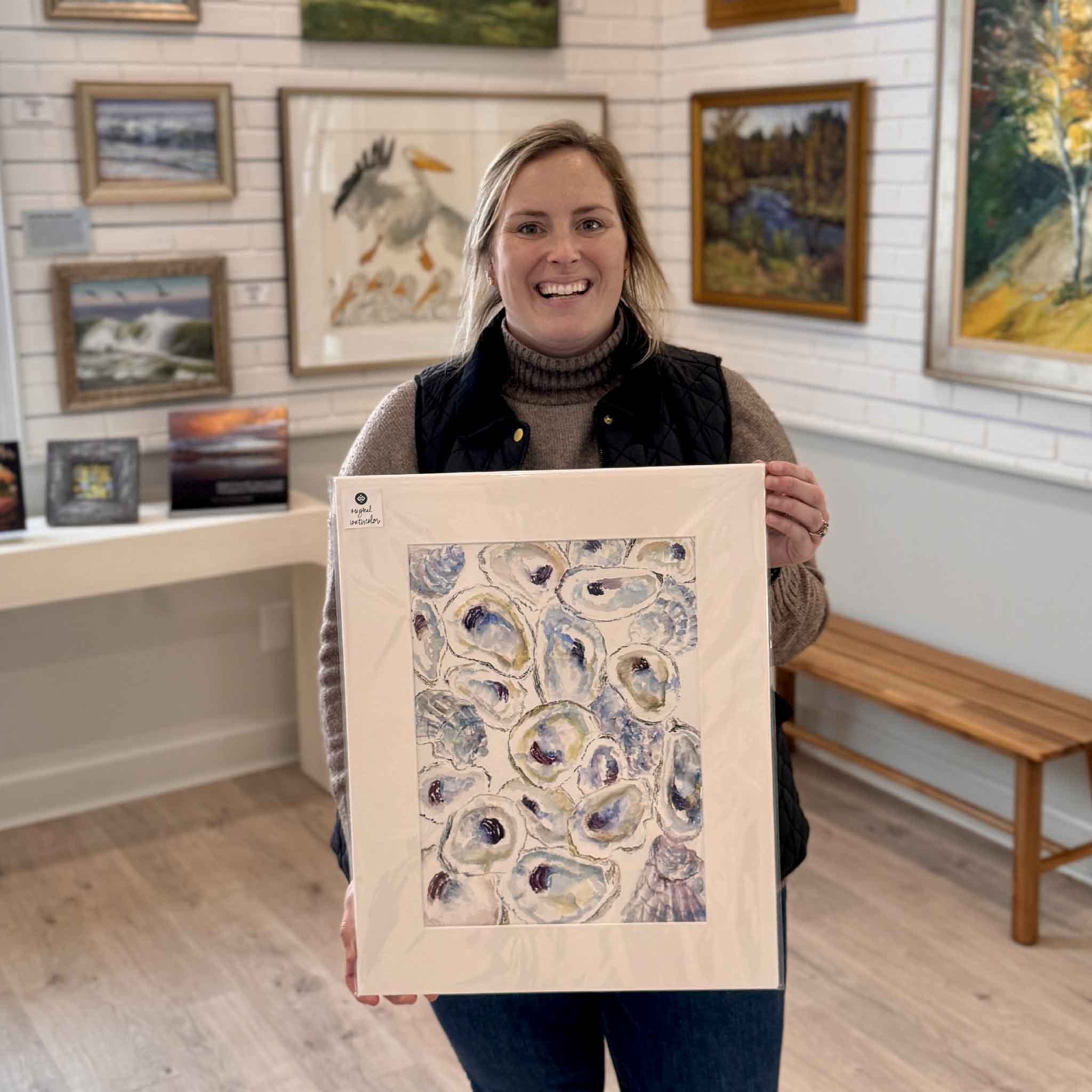 Gray Man Gallery team member holding Gathered Jewels, an original oyster watercolor by local artist Allison Farr, matted 16x20 and displayed inside the gallery in Pawleys Island