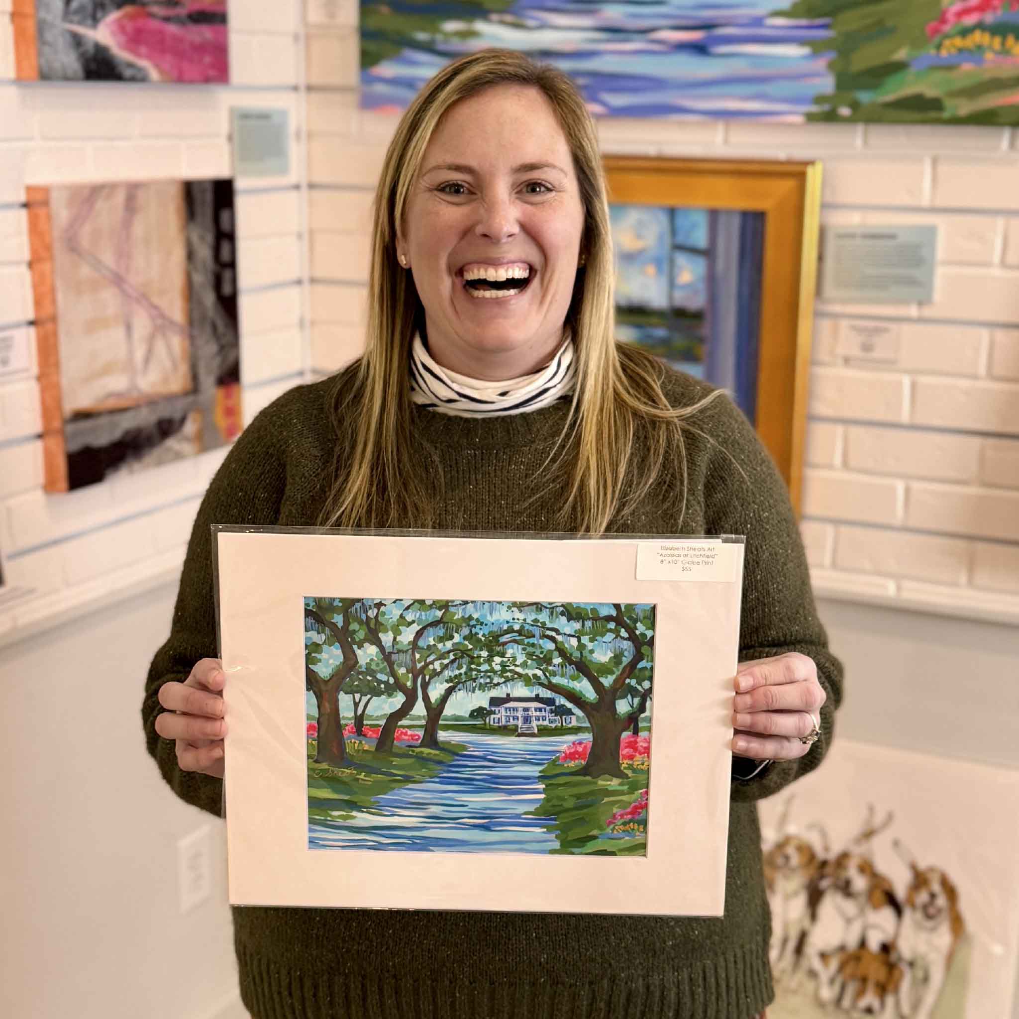 Elizabeth Sheats Azaleas at Litchfield 11x14 matted fine art giclée print held by staff inside Gray Man Gallery in Pawleys Island South Carolina, featuring Litchfield Plantation entrance with live oaks, Spanish moss, pink azaleas, and classic Lowcountry coastal landscape artwork