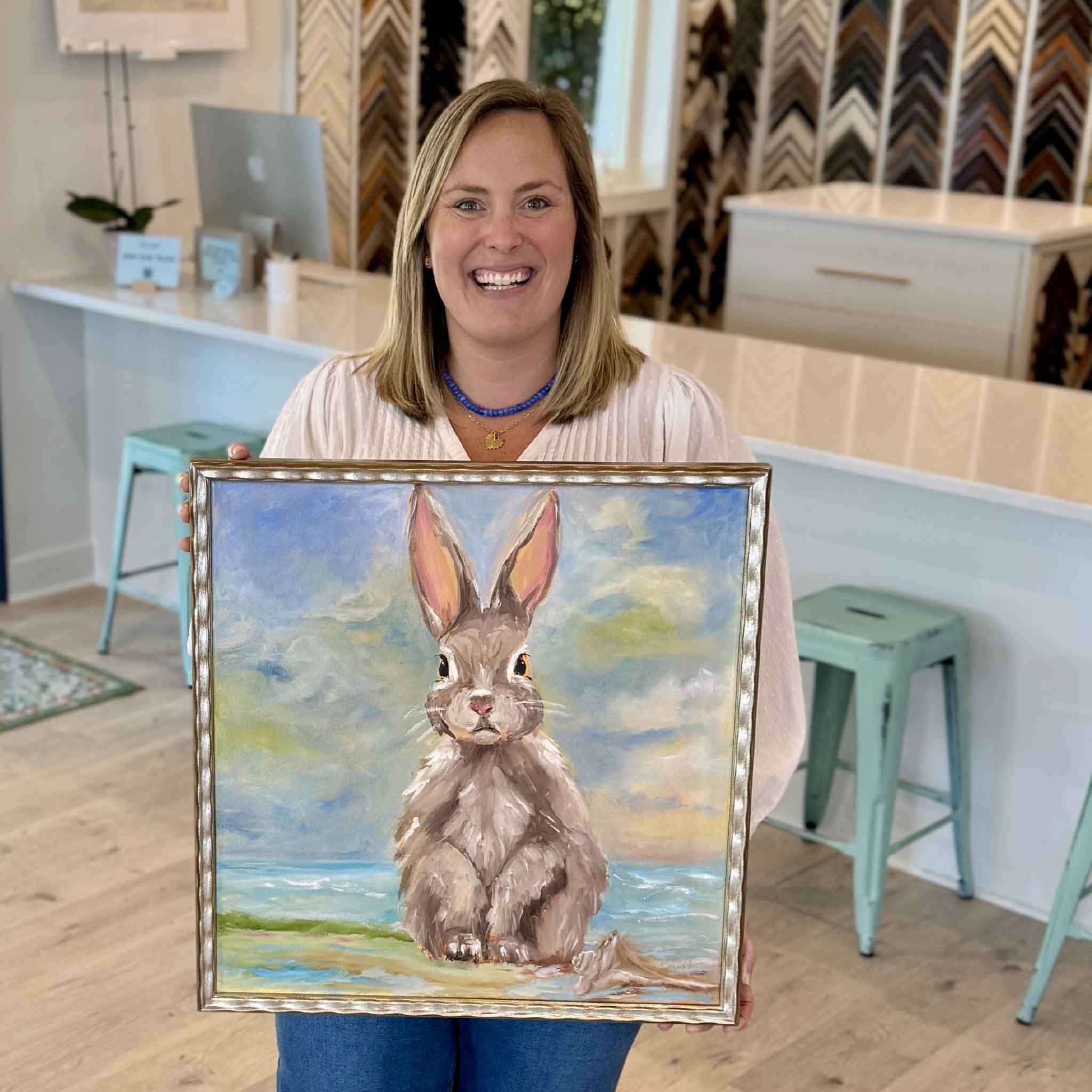 Beach Bunny original oil painting by local artist Jenny Reynolds featuring a rabbit seated on a sandy shoreline with ocean waves and a soft pastel sky behind it framed in her signature champagne frame at Gray Man Gallery