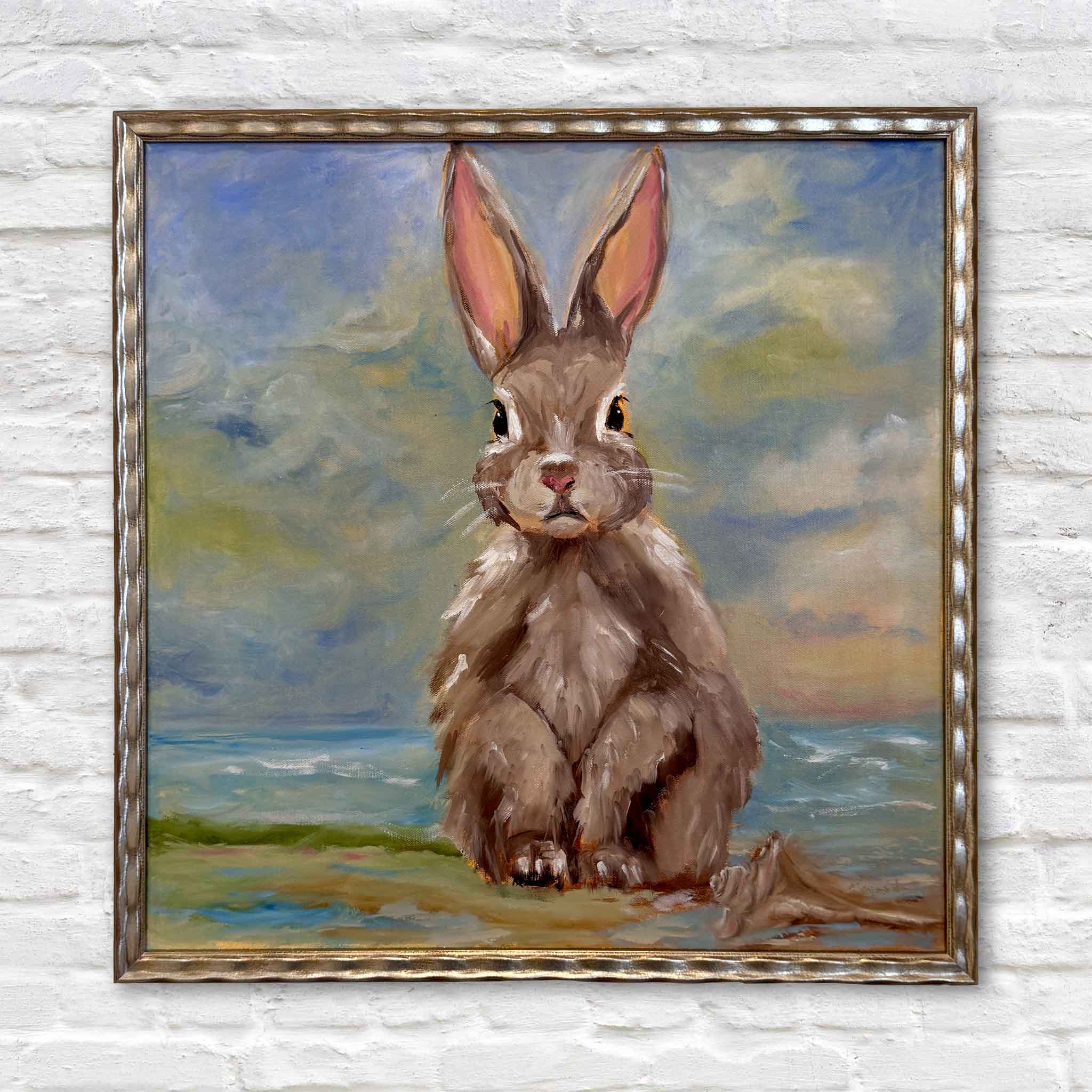 Bordy holding Beach Bunny by local artist Jenny Reynolds inside Gray Man Gallery Pawleys Island showing scale of original oil painting of a rabbit sitting on sand with ocean waves and soft coastal sky in a champagne frame