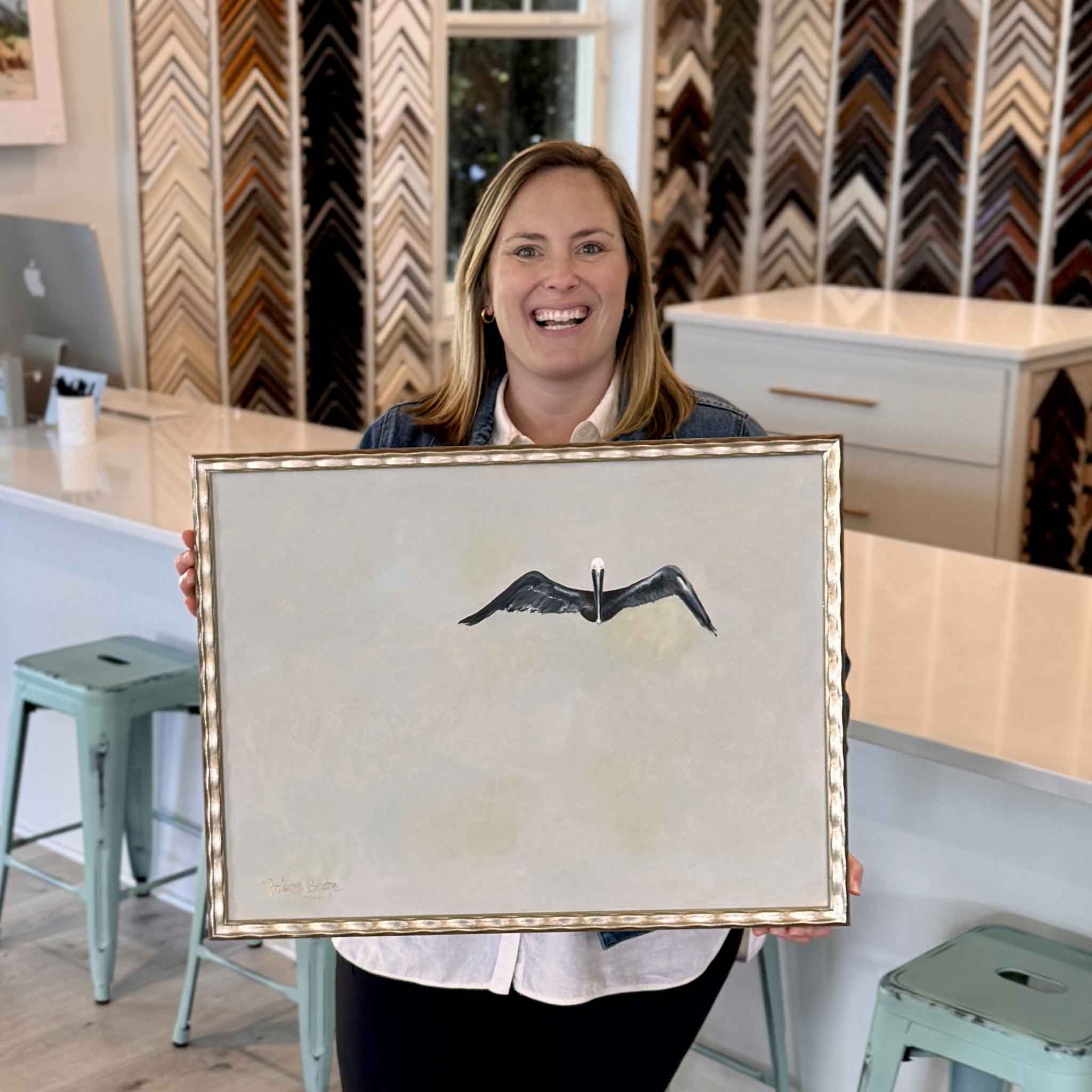 Bordy holding One by Braden Grafe inside Gray Man Gallery in Pawleys Island, framed oil painting of a single pelican in flight with expansive negative space and soft neutral palette, showcasing scale and detail of this minimal Lowcountry coastal artwork