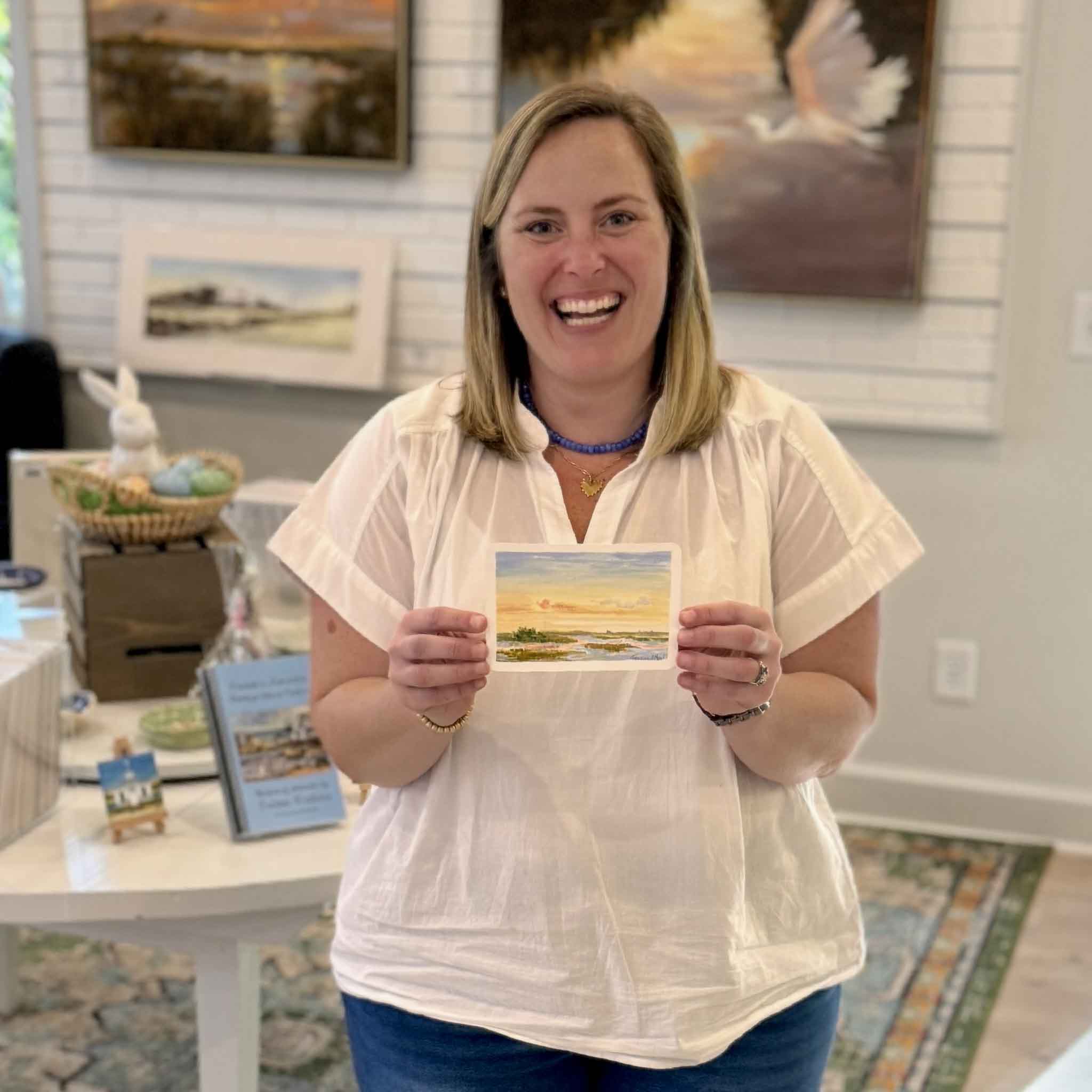 Bordy holding First Light by Jeanne O’Neal inside Gray Man Gallery in Pawleys Island showing the scale of the 6x4 original gouache painting on postcard with soft pastel sky and calm coastal water