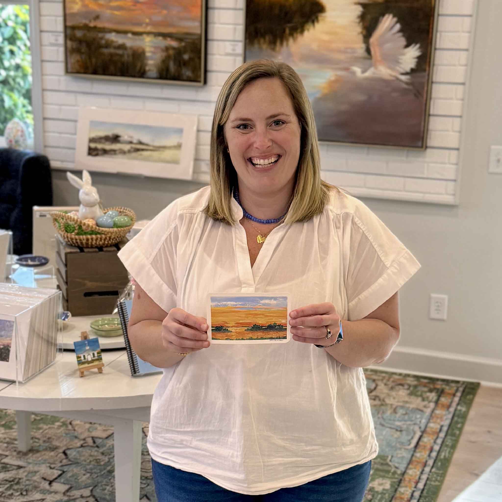 Bordy holding Island Glow by Jeanne O’Neal inside Gray Man Gallery in Pawleys Island showing the scale of the 6x4 original gouache sunset painted on postcard with glowing horizon and soft island silhouettes