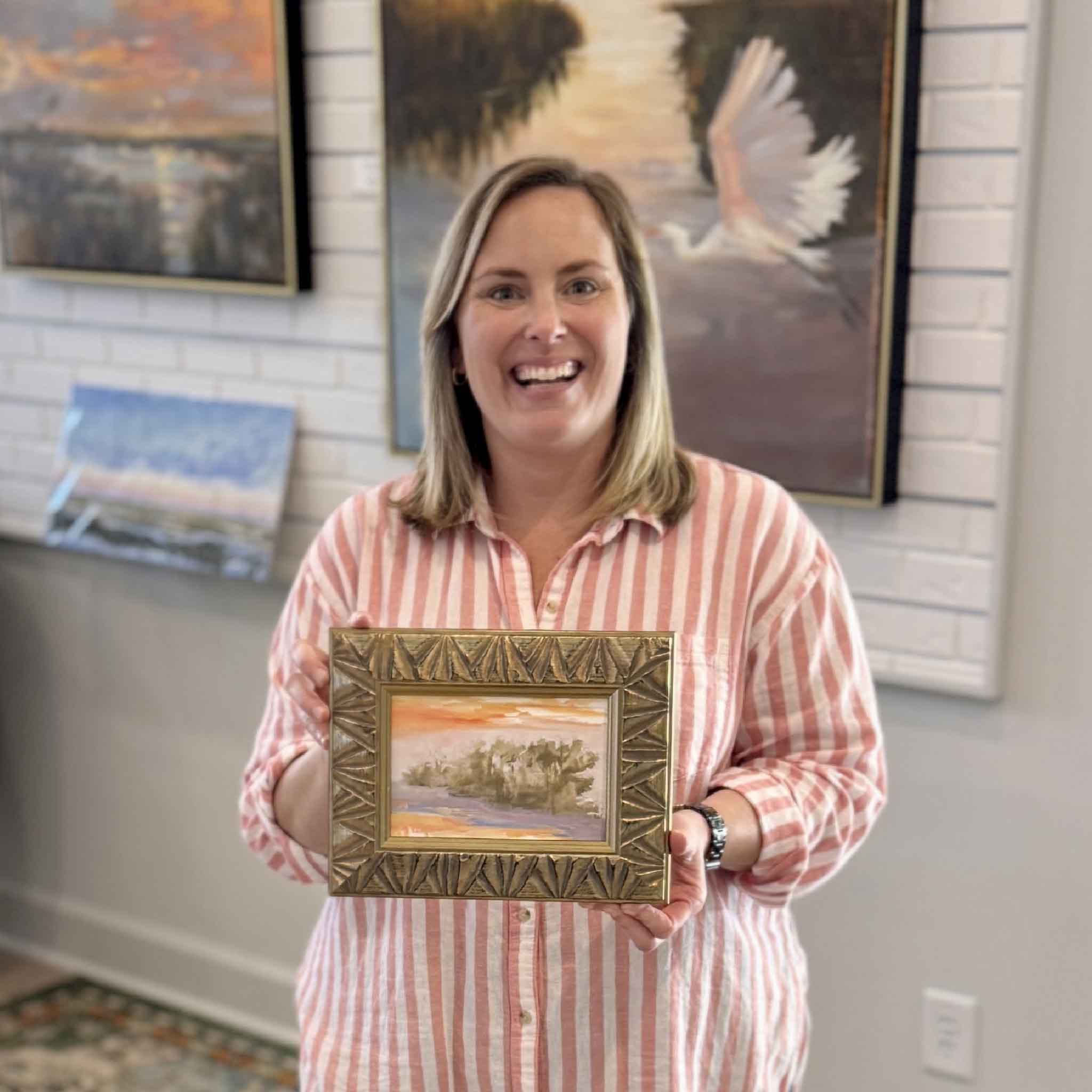 Bordy holding Edge of the Tide by Jeanne O’Neal inside Gray Man Gallery in Pawleys Island, small original oil painting with warm sunset tones, marsh landscape, and ornate gold frame for scale and presentation