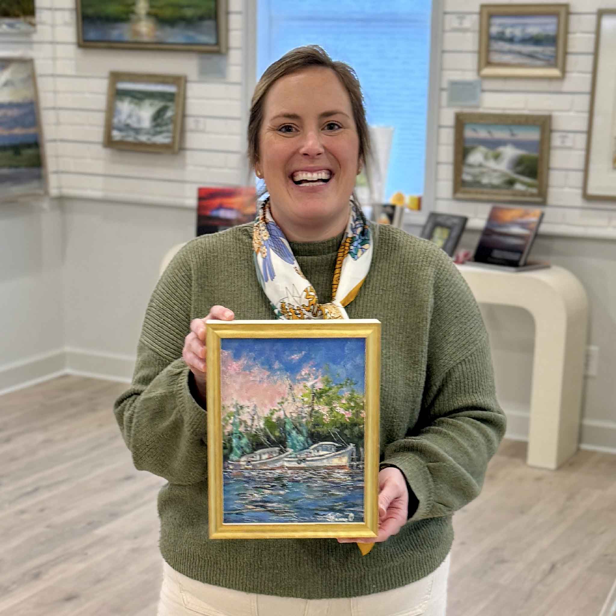 Bordy of Gray Man Gallery standing inside the Pawleys Island gallery holding “Evening Light,” an original oil painting by Jeanne O’Neal depicting shrimp boats at sunset on the Georgetown South Carolina waterfront, showcasing the scale, framing craftsmanship, and curated coastal art environment of the Lowcountry’s trusted local art gallery