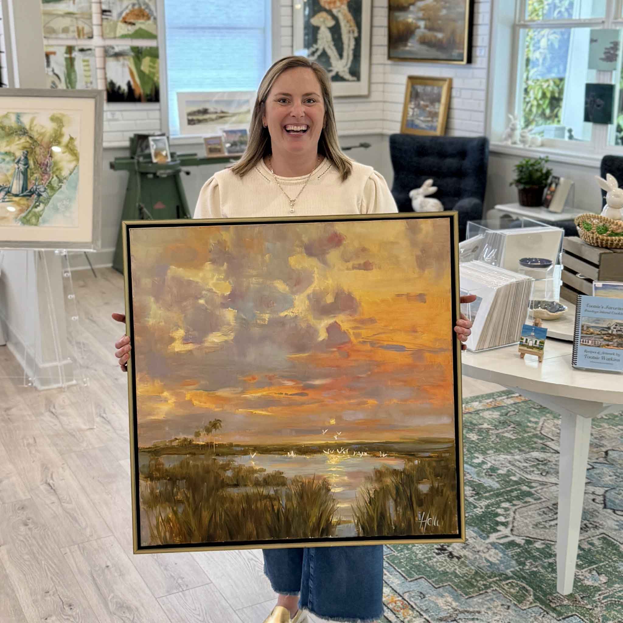 Bordy holding Nature’s Splendor by local artist Helli Luck inside Gray Man Gallery Pawleys Island showing scale of framed Lowcountry sunset painting