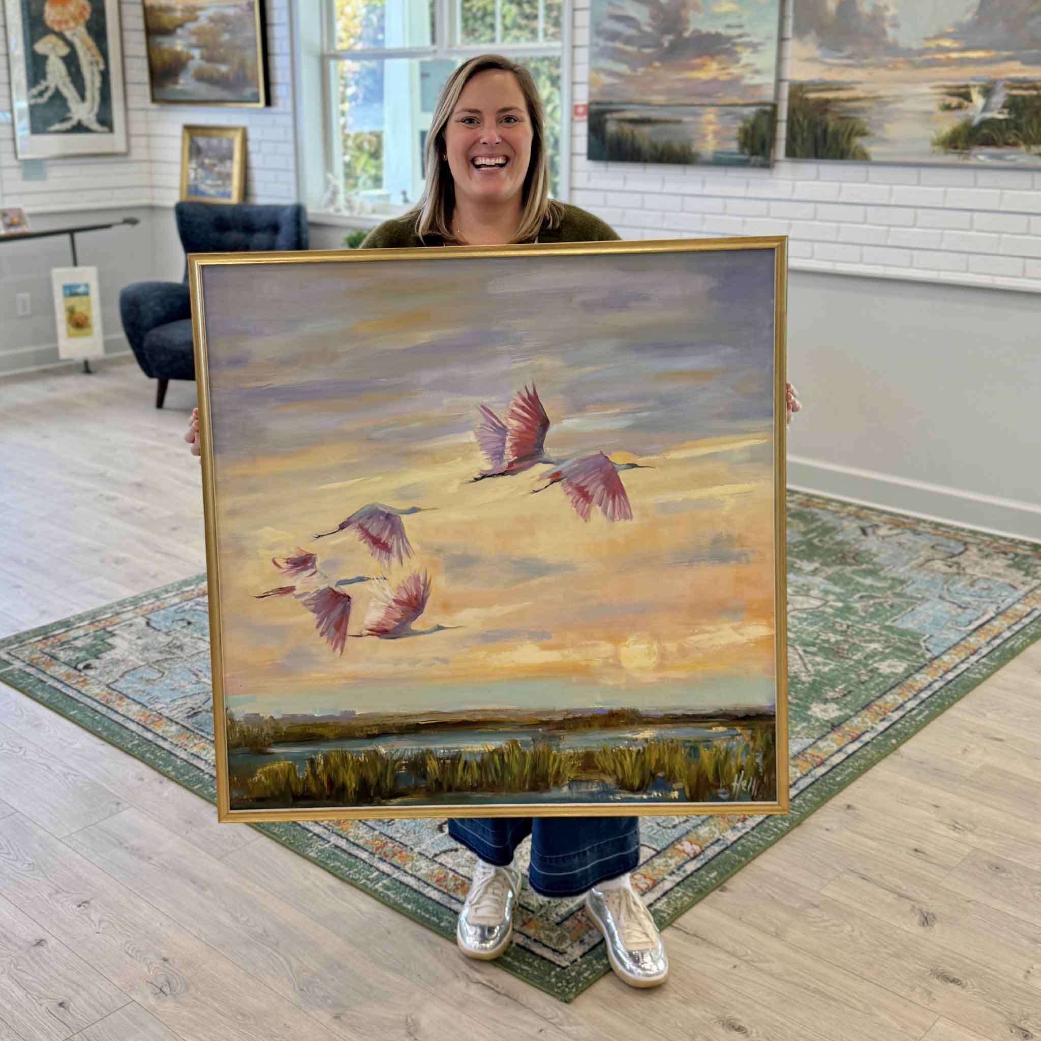 Bordy holding Twilight Ballet original painting by Helli Luck inside Gray Man Gallery Pawleys Island, showing scale of the 36 by 36 inch framed marsh scene with roseate spoonbills at sunset