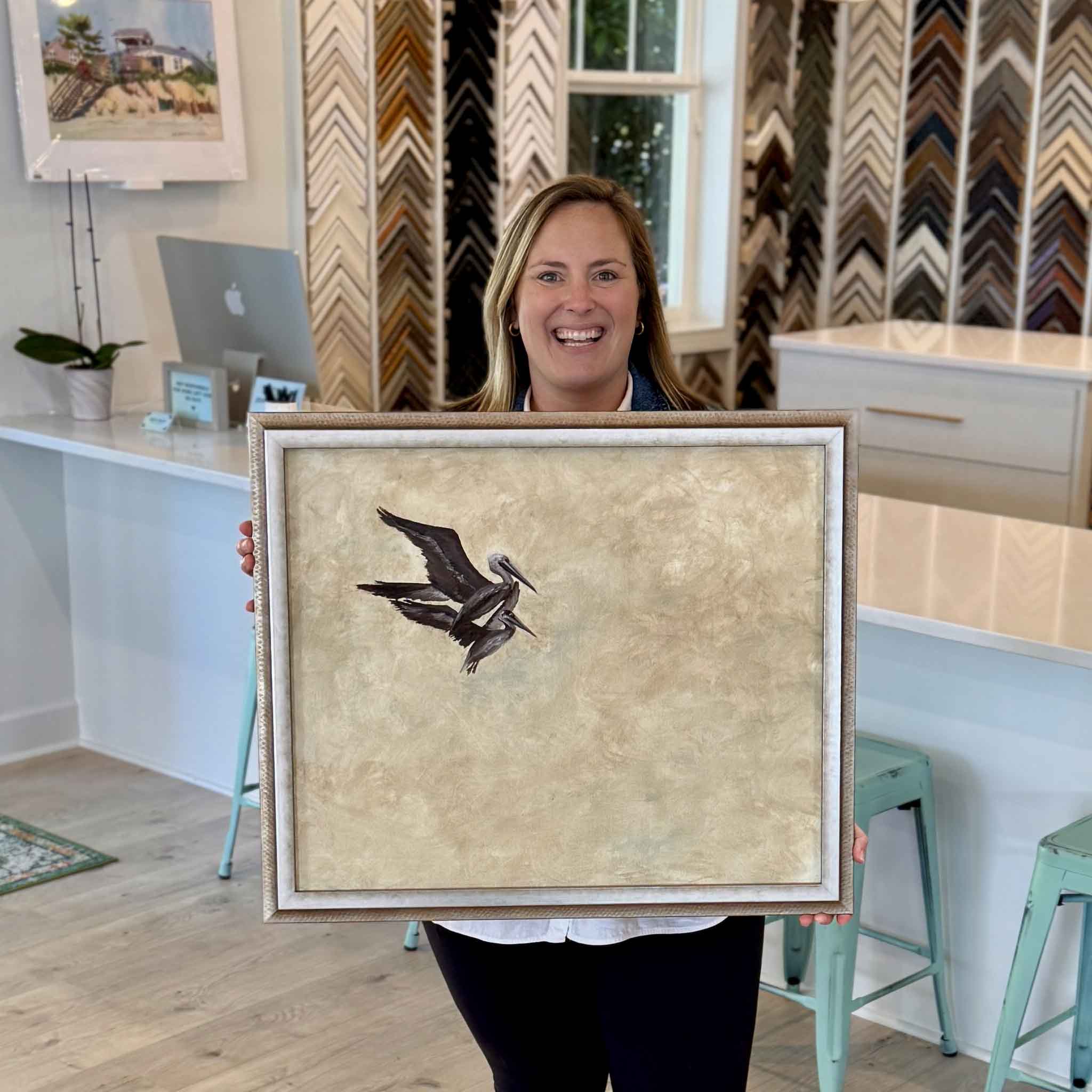 Bordy holding Flight Study I by Braden Grafe inside Gray Man Gallery in Pawleys Island, framed oil painting of two pelicans in flight with a subtle neutral palette and textured background coastal artwork by local artist