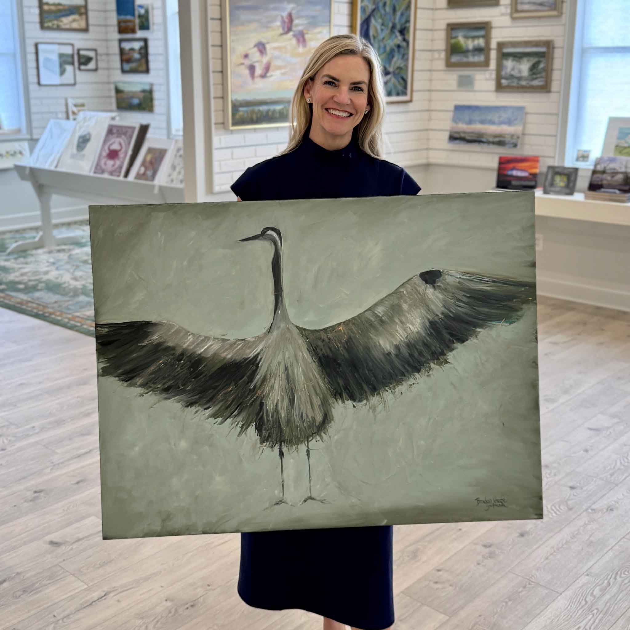 Local artist Braden Grafe holding her painting Wings of the Dawn inside Gray Man Gallery in Pawleys Island, gallery wrapped oil painting of a great blue heron with expansive wingspan, neutral coastal palette, and textured painterly surface