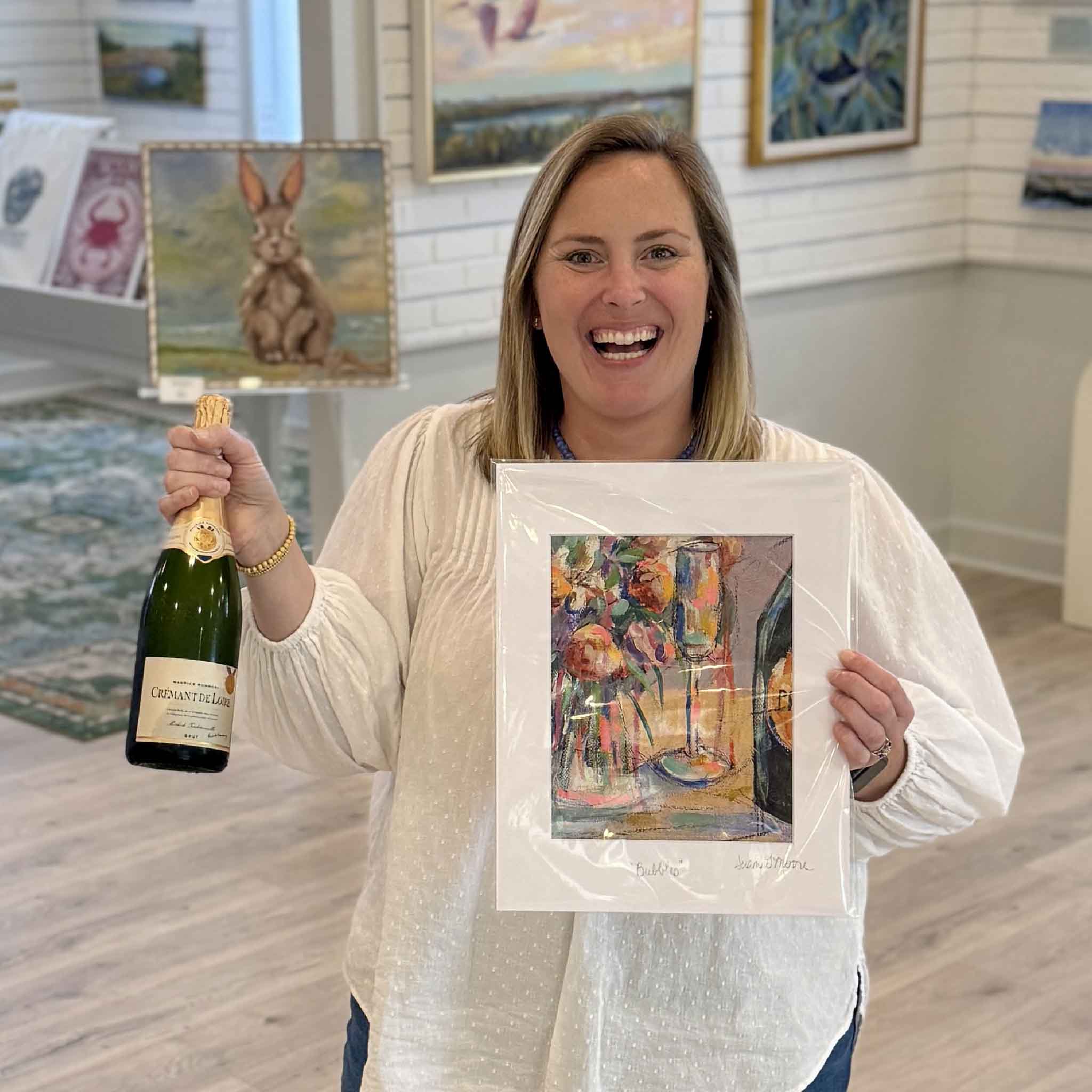Bordy holding Bubbles by local artist Susan Moore at Gray Man Gallery Pawleys Island alongside a champagne bottle showing scale of colorful giclee print featuring a champagne glass and floral bouquet with expressive brushwork
