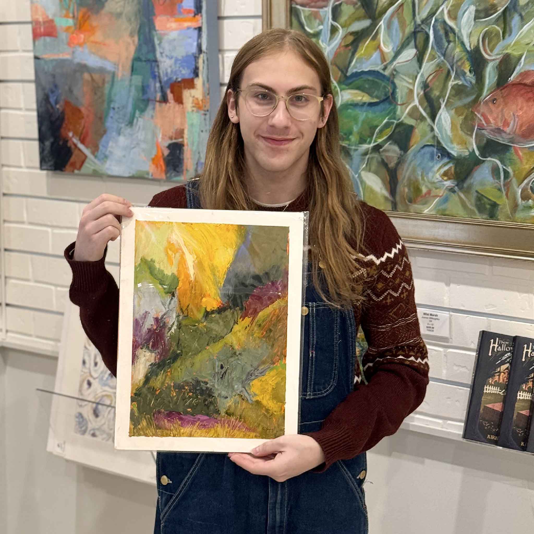 Alex holding abstract mixed media painting “Into the Thicket” by Caroline Young featuring bold textured strokes in green, gold, violet, and rust, evoking a wild forest landscape.