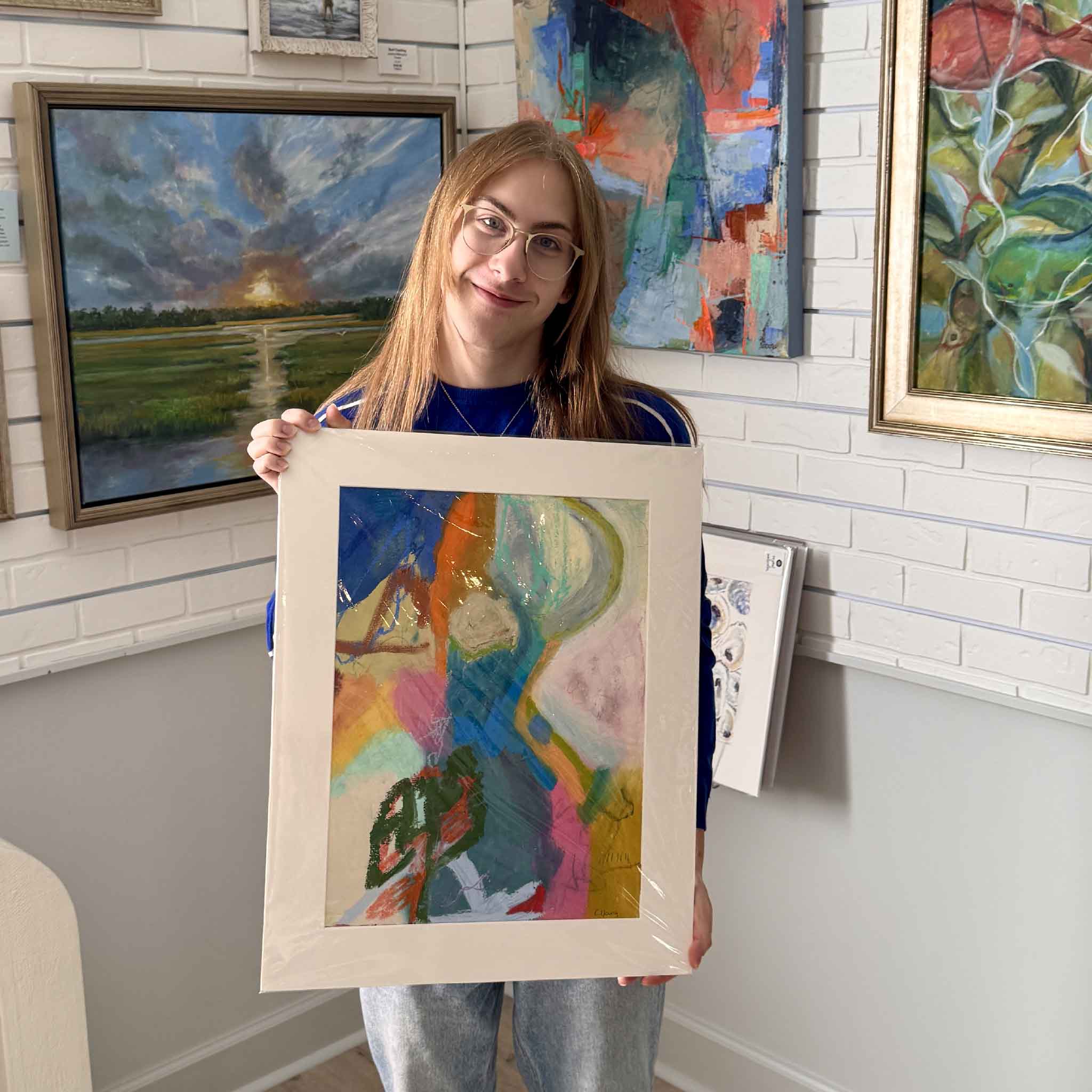 Alex holding the original mixed-media painting Riverbend by Caroline Young, a vibrant abstract landscape in saturated blues, pinks, and greens, shown at Gray Man Gallery.