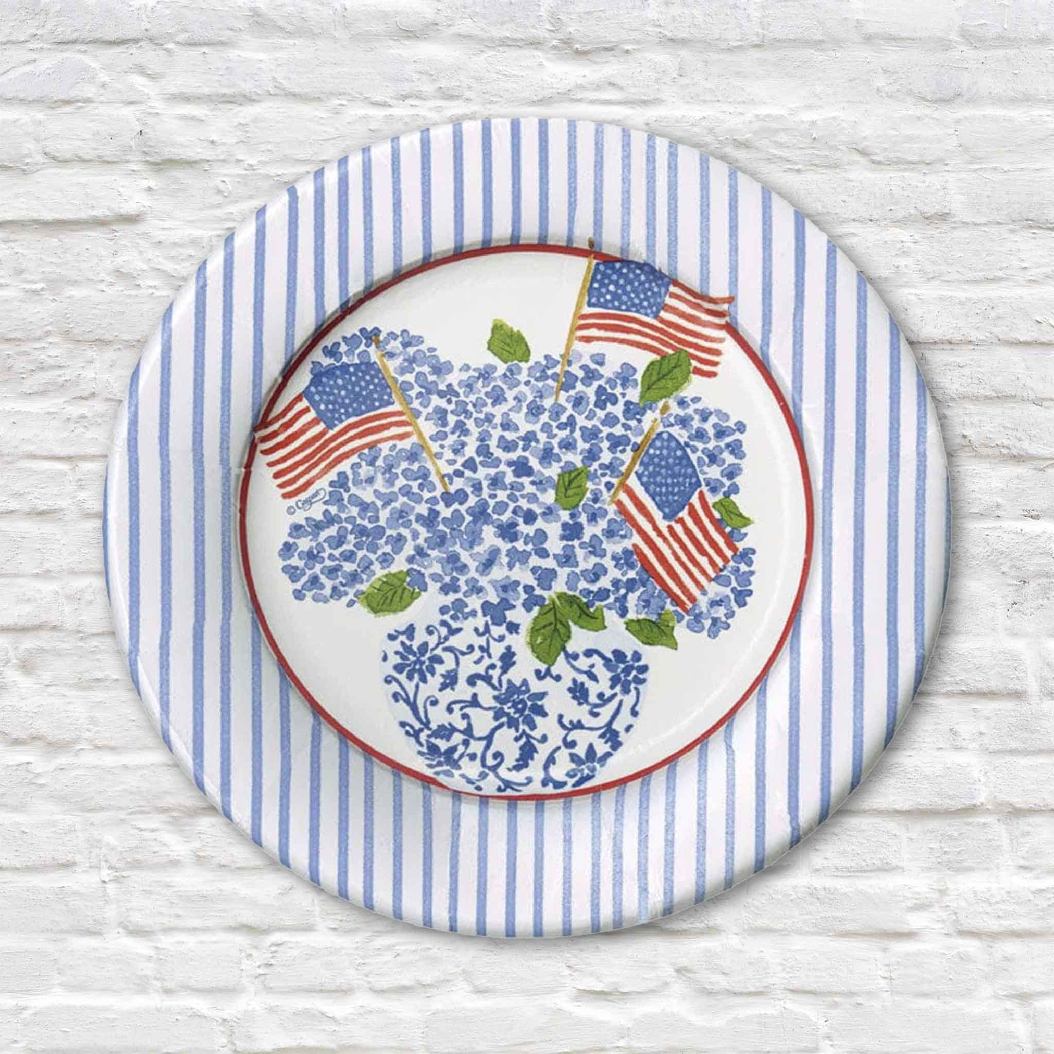 A Caspari Americana Flags & Hydrangeas 8-inch salad and dessert plate featuring Lindsay Brackeen's artwork of blue hydrangeas arranged in a blue-and-white chinoiserie vase with waving American flags, surrounded by a classic blue striped border and red accent rim. These premium plastic-coated paper plates are made in the USA and sold in packs of eight at Gray Man Gallery in Pawleys Island, South Carolina.