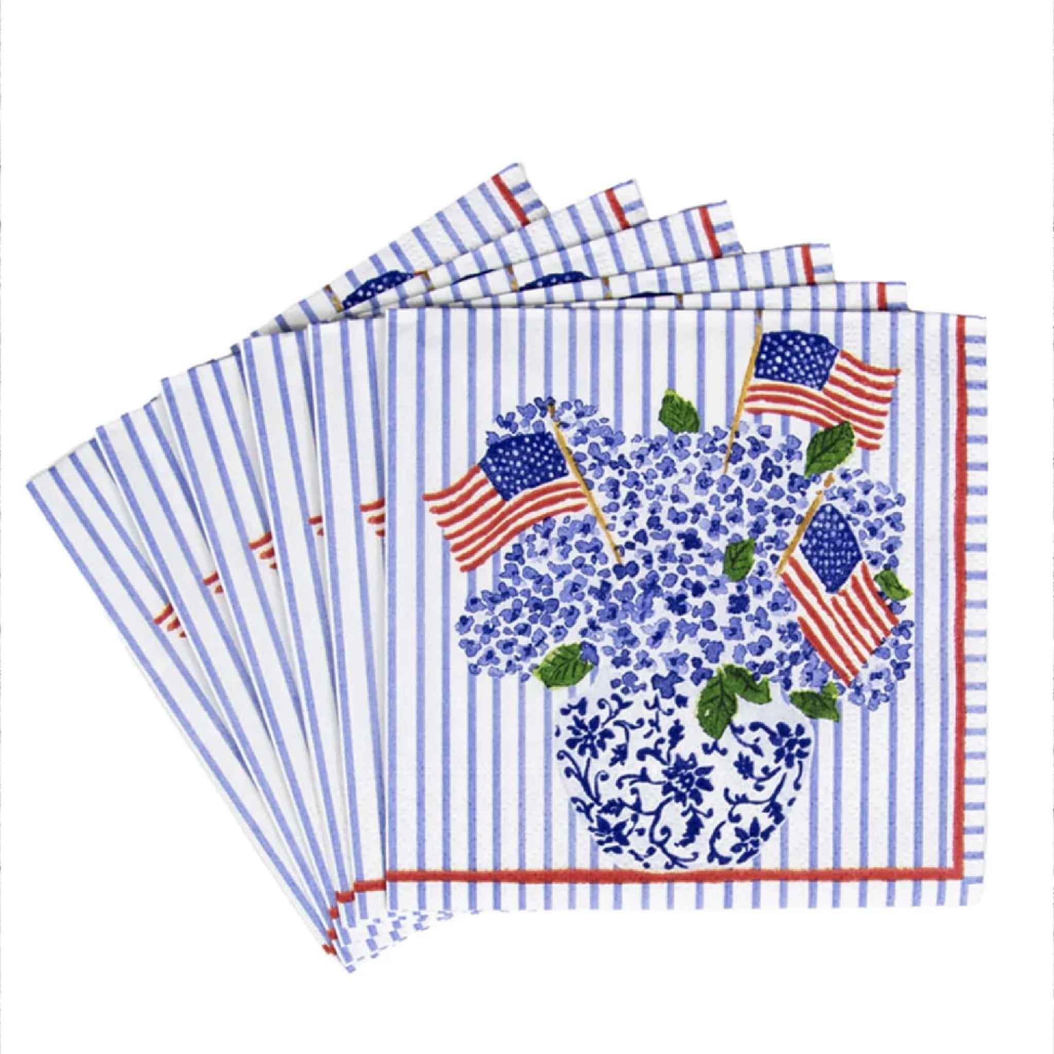 A fan of Caspari Flags & Hydrangeas cocktail napkins by Lindsay Brackeen displays the complete set of 20 premium triple-ply paper napkins featuring blue hydrangeas, American flags, and a blue-and-white chinoiserie vase. Available at Gray Man Gallery in Pawleys Island, South Carolina for patriotic celebrations and coastal entertaining.