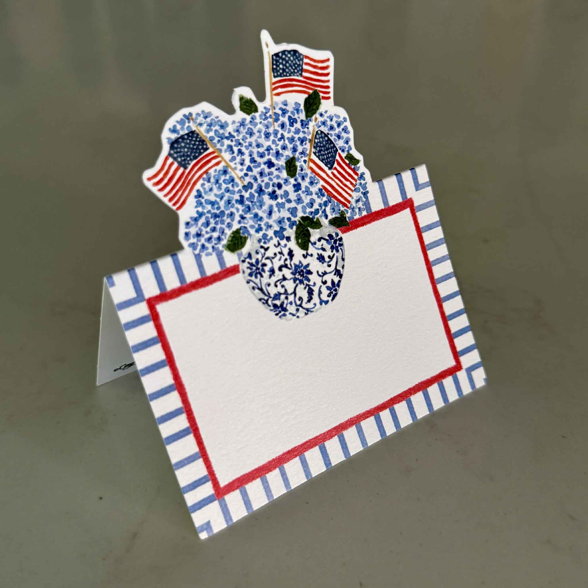 Caspari Flags & Hydrangeas die-cut tented place card featuring artwork by Lindsay Brackeen, showcasing a blue-and-white chinoiserie vase overflowing with blue hydrangeas and accented by American flags above a generously sized writing area with a red border and blue striped frame. Available at Gray Man Gallery in Pawleys Island, South Carolina, these premium cardstock place cards are sold as a set of eight and are perfect for Fourth of July celebrations, Memorial Day, Labor Day, entertaining, coastal dinners