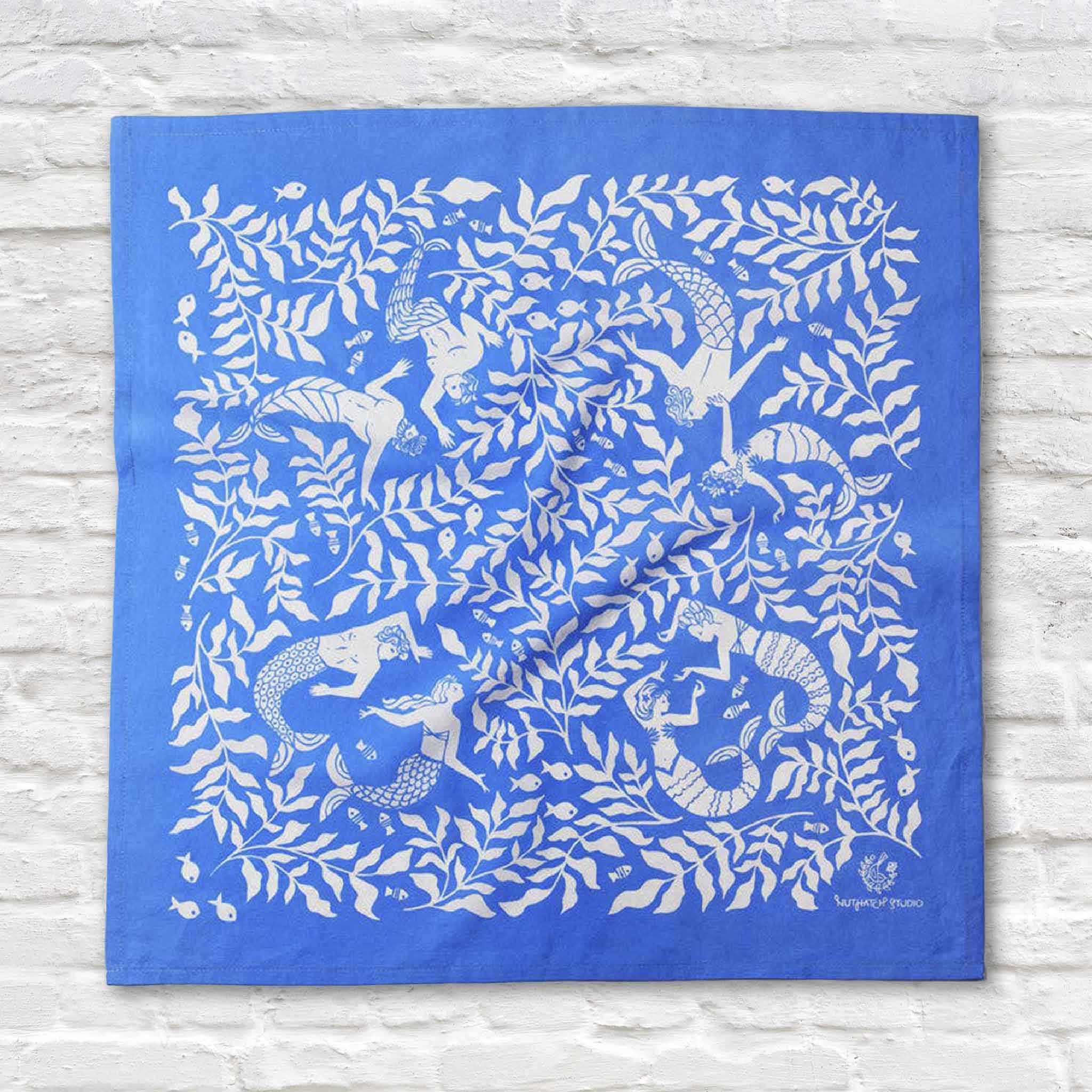 Cerulean Mermaid organic cotton bandana by Chrissie Van Hoever displayed flat at Gray Man Gallery in Pawleys Island, South Carolina. The 22-inch square textile features elegant white mermaids swimming through flowing kelp forests on a vibrant cerulean blue background, making it beautiful to wear as a scarf or display as framed coastal wall art.