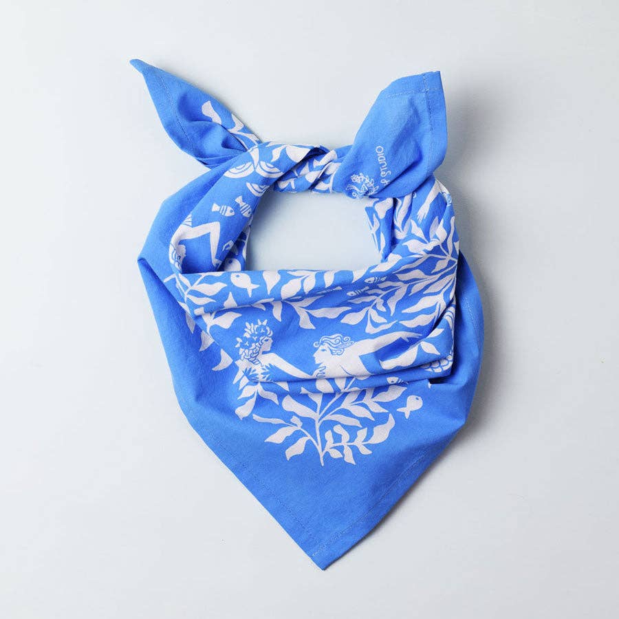 Cerulean Mermaid organic cotton bandana by Chrissie Van Hoever styled as a neck scarf at Gray Man Gallery in Pawleys Island, South Carolina. This 22-inch square bandana features whimsical white mermaids swimming through flowing kelp forests on a vibrant cerulean blue background.