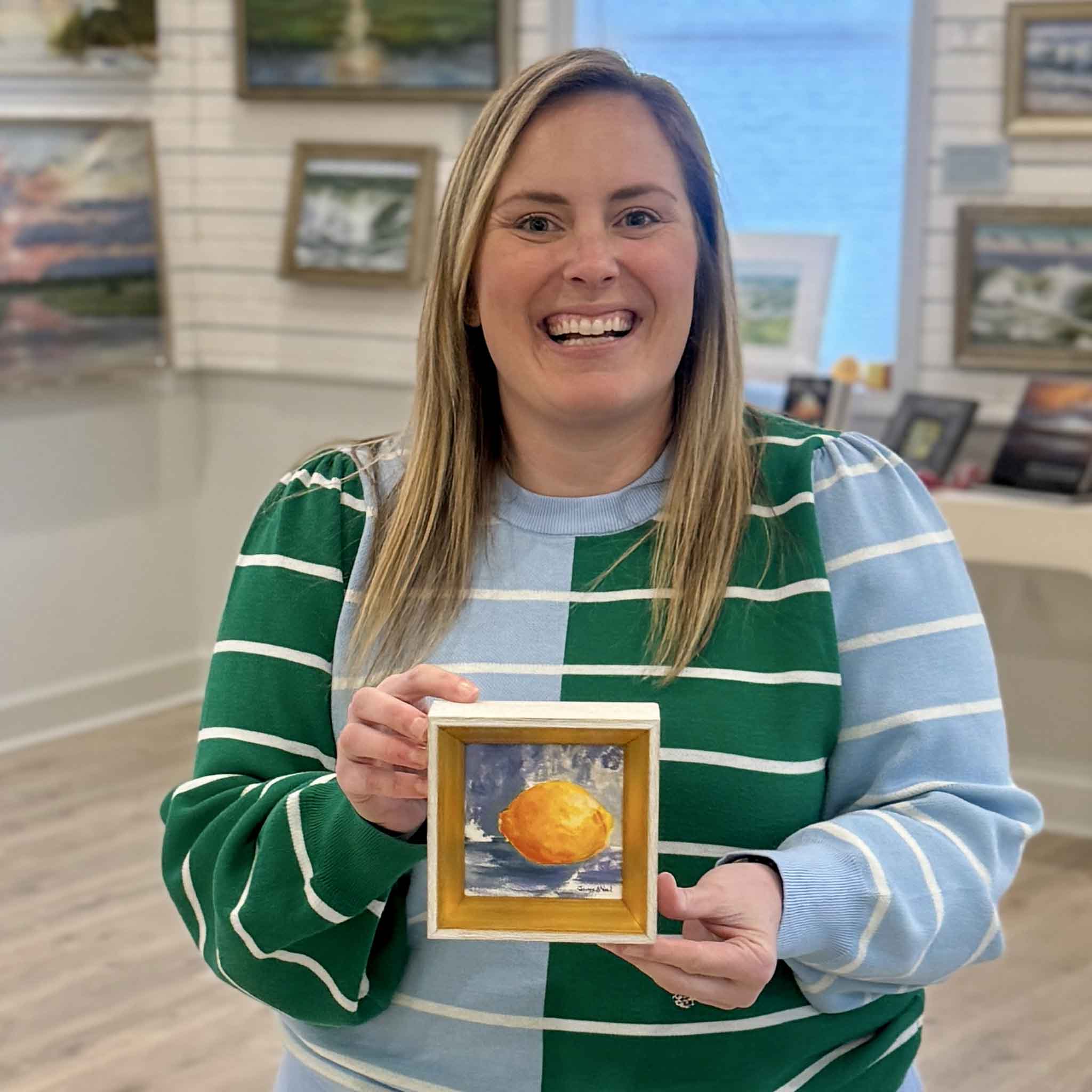 Bordy holds “Citrus Light,” an original 4x4 oil painting by Jeanne O’Neal, inside Gray Man Gallery in Pawleys Island, South Carolina. The image highlights the scale, signature gold frame, and luminous citrus brushwork of this collectible Lowcountry still life artwork.