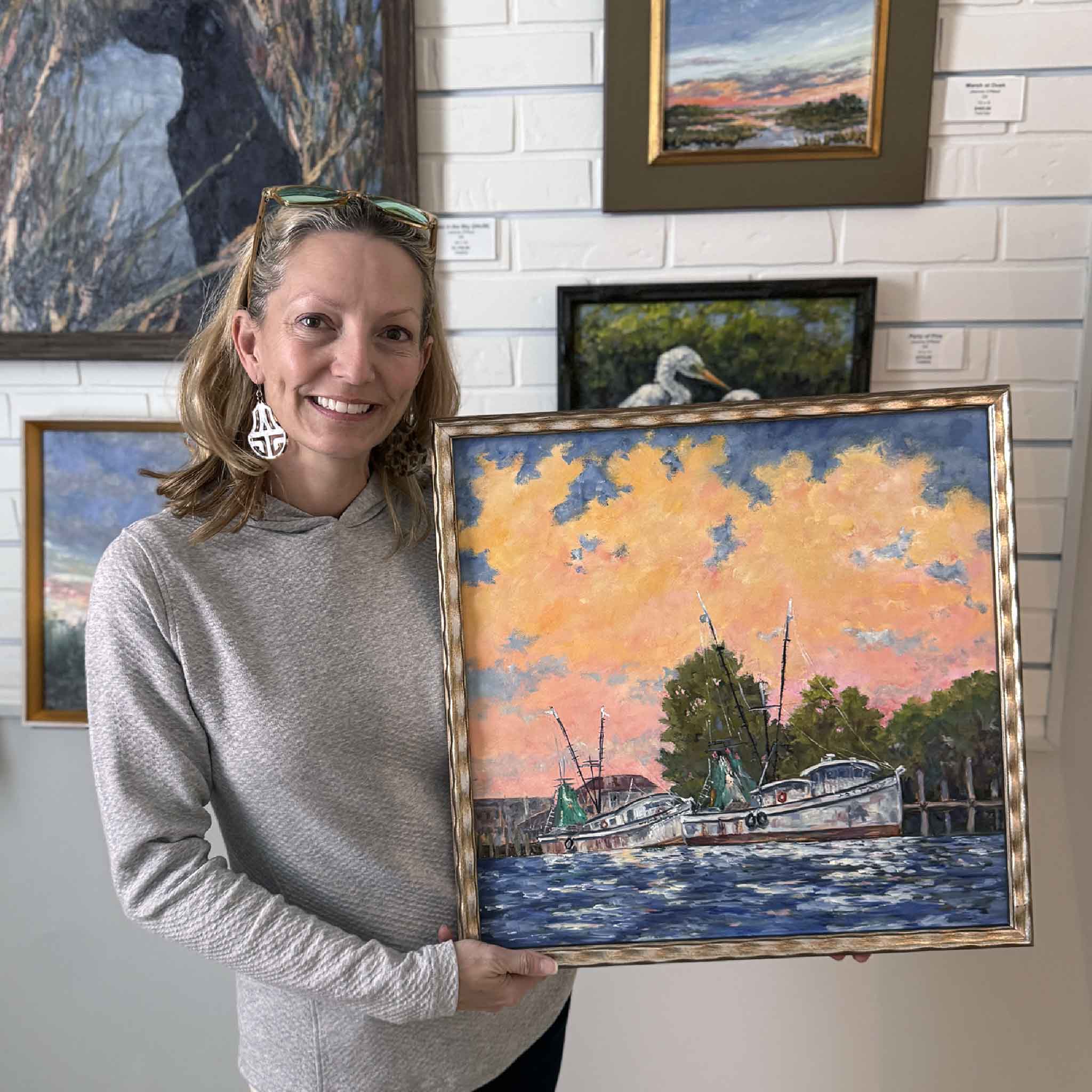 Local artist Jeanne O’Neal holding her framed painting “Dockside Light,” showing vibrant shrimp boats at sunset inside Gray Man Gallery.