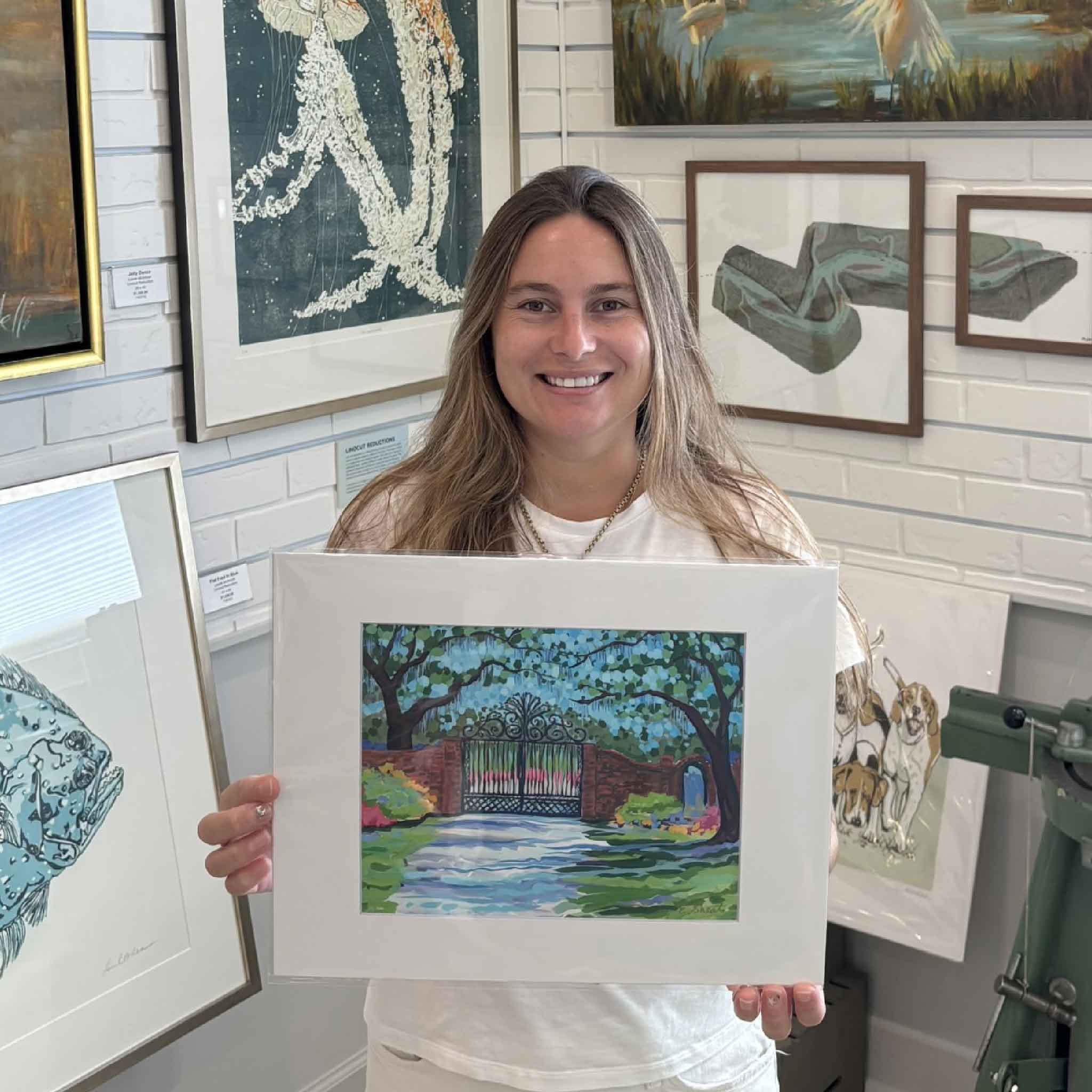 Kyra holding the smaller 11x14 matted Litchfield Gate fine art print by Elizabeth Sheats inside Gray Man Gallery in Pawleys Island, South Carolina, a colorful Lowcountry artwork of the historic Litchfield Plantation gates sized to fit a standard frame and ideal for easy gifting.