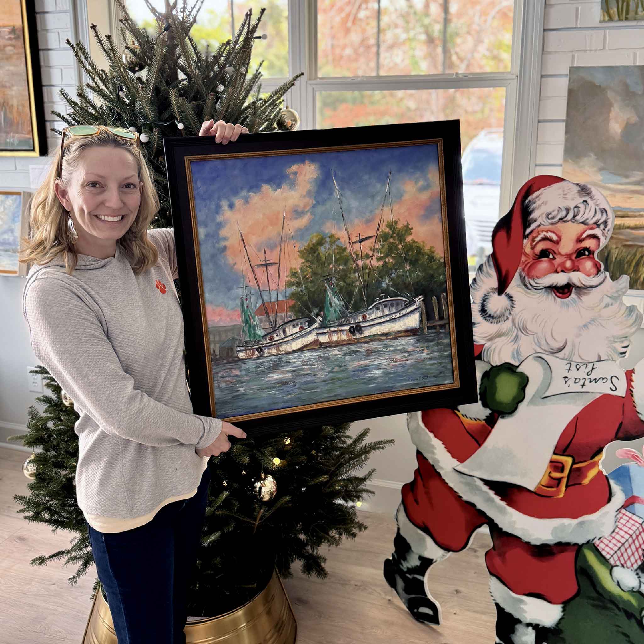 Artist Jeanne O’Neal holding her framed oil painting “End of Day” inside Gray Man Gallery, shown for scale near a Christmas display.
