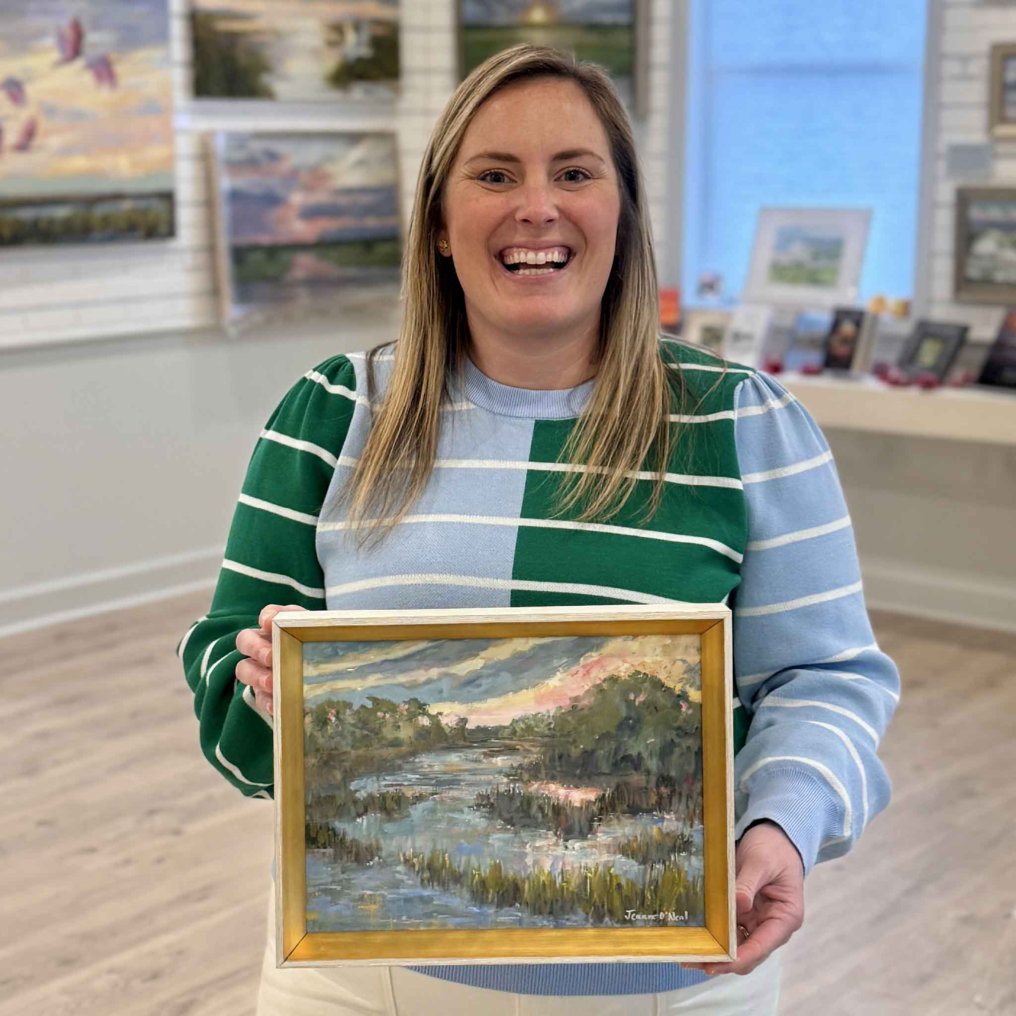 Bordy holding End of Summer, an original oil painting by Jeanne O’Neal in a warm gold frame, featuring a South Carolina Lowcountry marsh at sunset, inside Gray Man Gallery in Pawleys Island SC