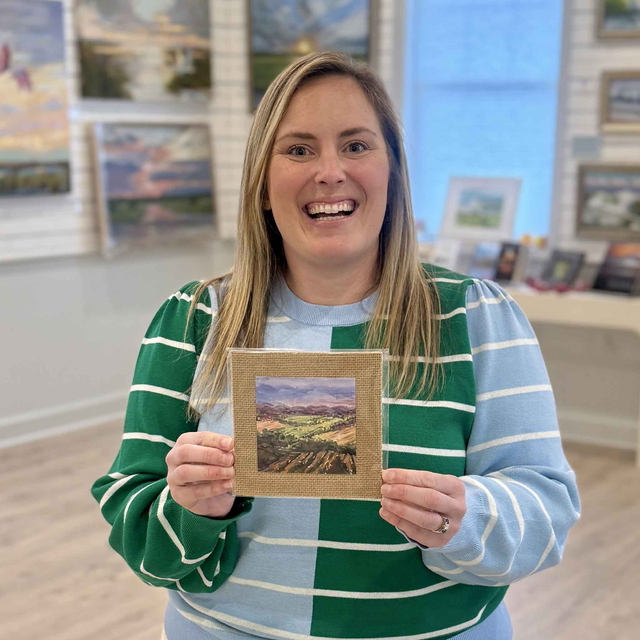 Bordy holds “Fields of France,” a 4x4 fine art print by Jeanne O’Neal, local artist represented at Gray Man Gallery in Pawleys Island, South Carolina. The image shows the scale of the miniature landscape print mounted on burlap matboard, highlighting its textured finish and giftable size.