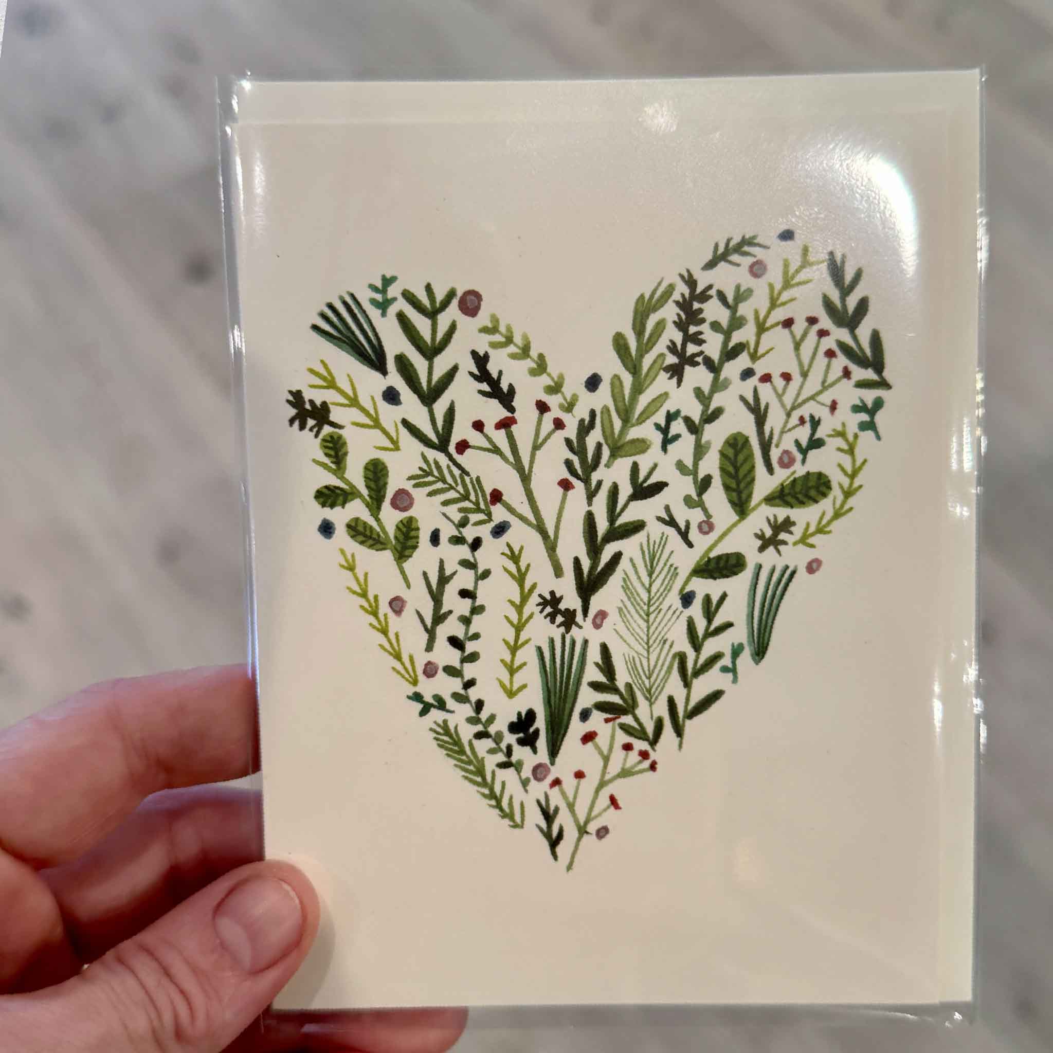 A hand holds the Floral Heart greeting card by Lizzy Stewart inside Gray Man Gallery in Pawleys Island, South Carolina. The elegant botanical illustration forms a heart from hand-painted greenery, wildflowers, berries, and delicate foliage, making this blank-inside greeting card a beautiful choice for anniversaries, weddings, Valentine's Day, sympathy, encouragement, or any heartfelt occasion.