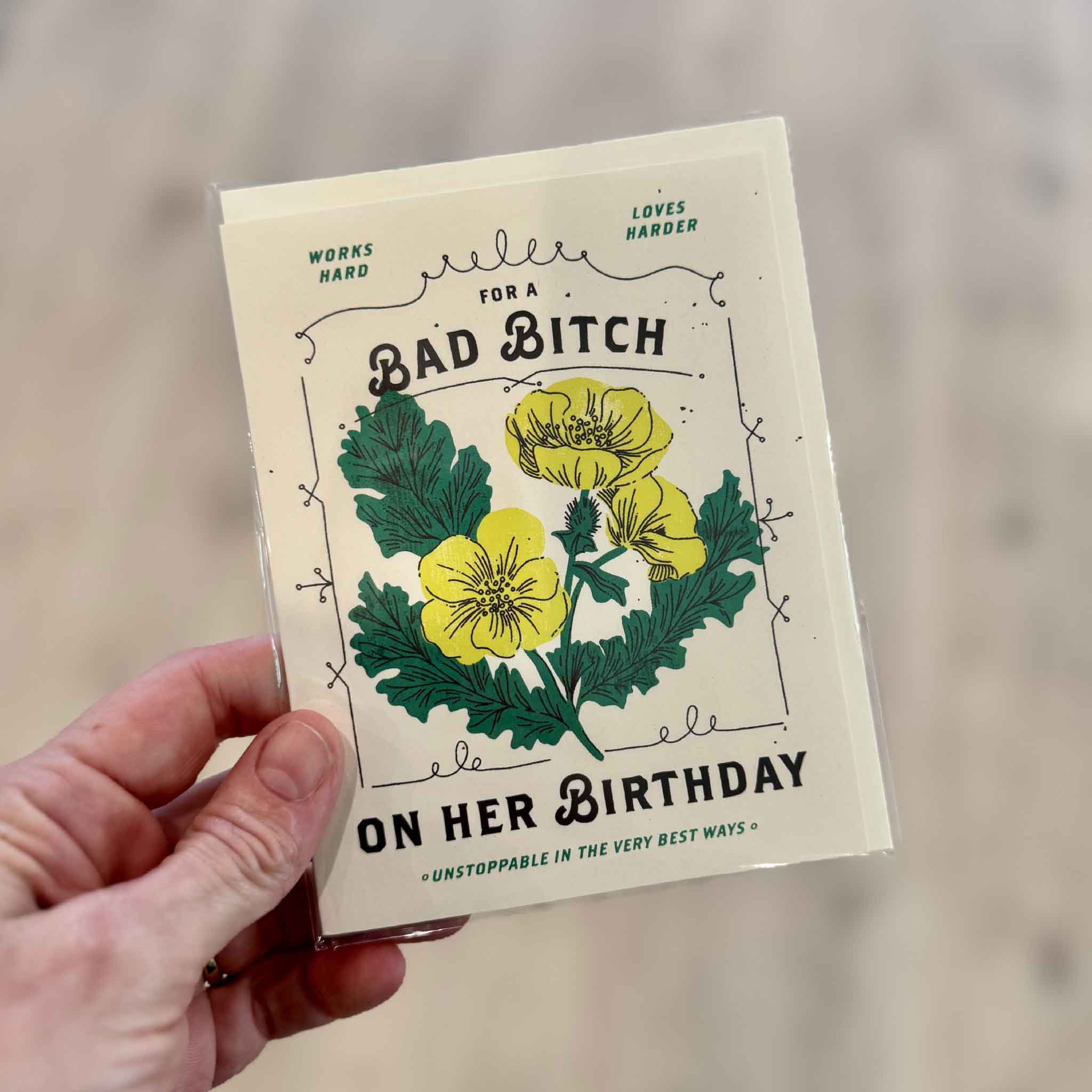 Hand holding the Bad Bitch Birthday Card, a funny floral birthday card for women with yellow flowers and the message “For a Bad Bitch on Her Birthday,” available at Gray Man Gallery in Pawleys Island, South Carolina.