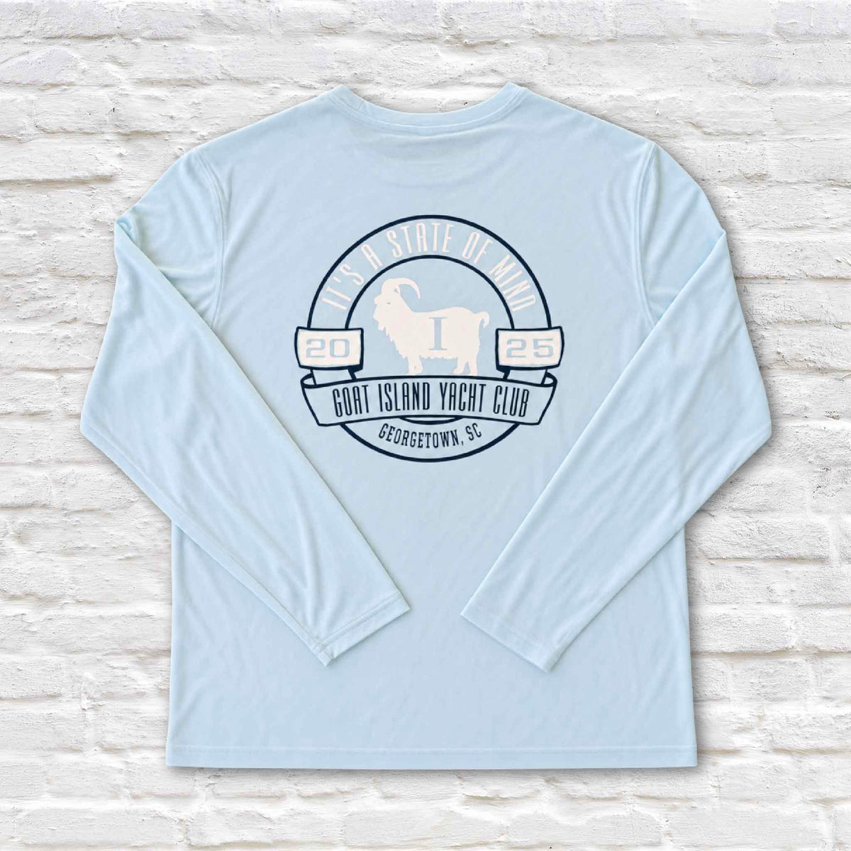 Back of a light blue Goat Island Yacht Club long sleeve performance fishing shirt featuring a large circular 2025 Georgetown, South Carolina logo with a white goat illustration and It's a State of Mind lettering. Made from 100% performance polyester by Altered Latitudes and photographed against the signature white brick background at Gray Man Gallery in Pawleys Island, South Carolina.