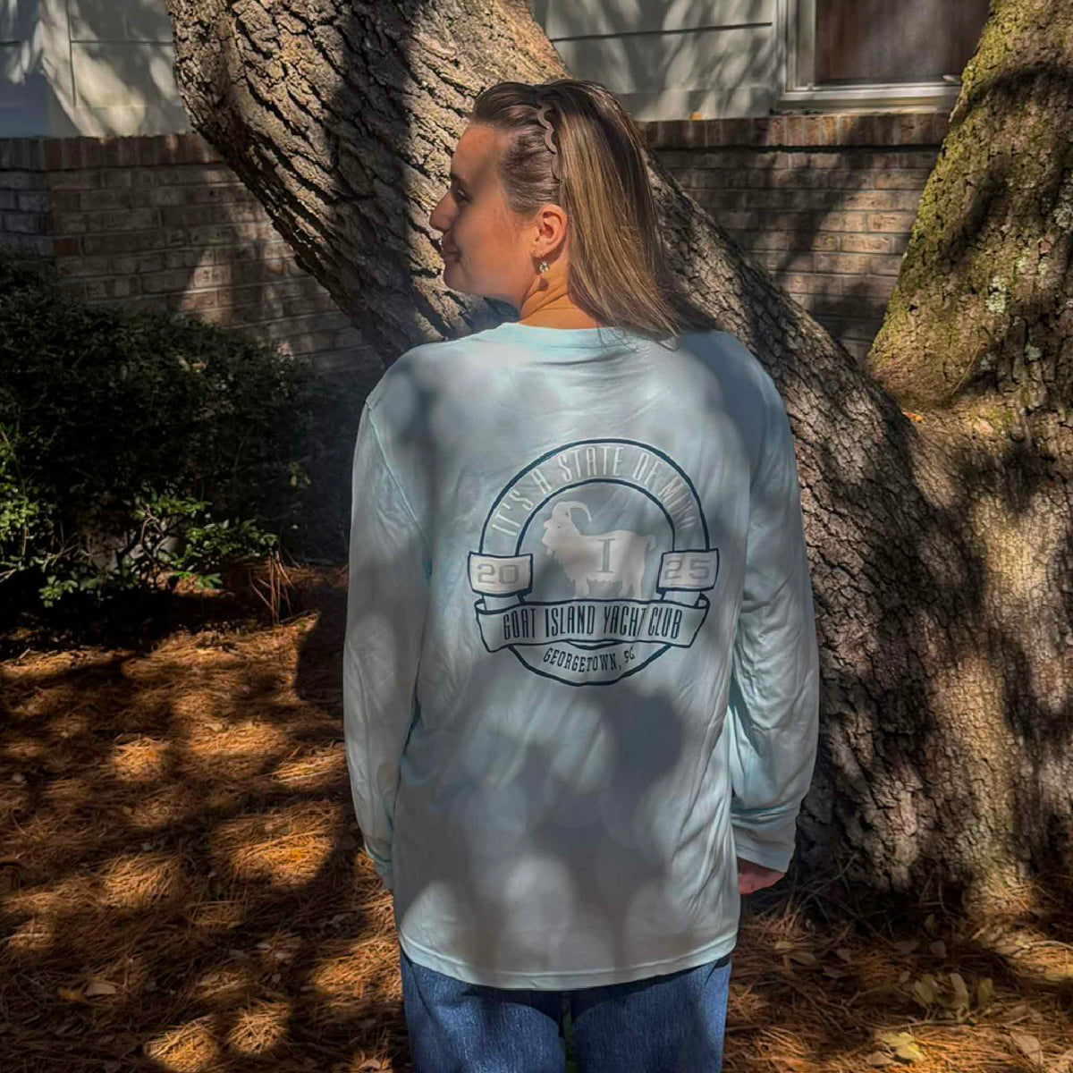 Kyra modeling the back of a light blue Goat Island Yacht Club long sleeve performance fishing shirt outdoors beneath a large oak tree in Pawleys Island, South Carolina. The back displays the Georgetown SC 2025 goat emblem and It's a State of Mind design, showing the shirt's relaxed fit and full-size nautical graphic. Available at Gray Man Gallery.