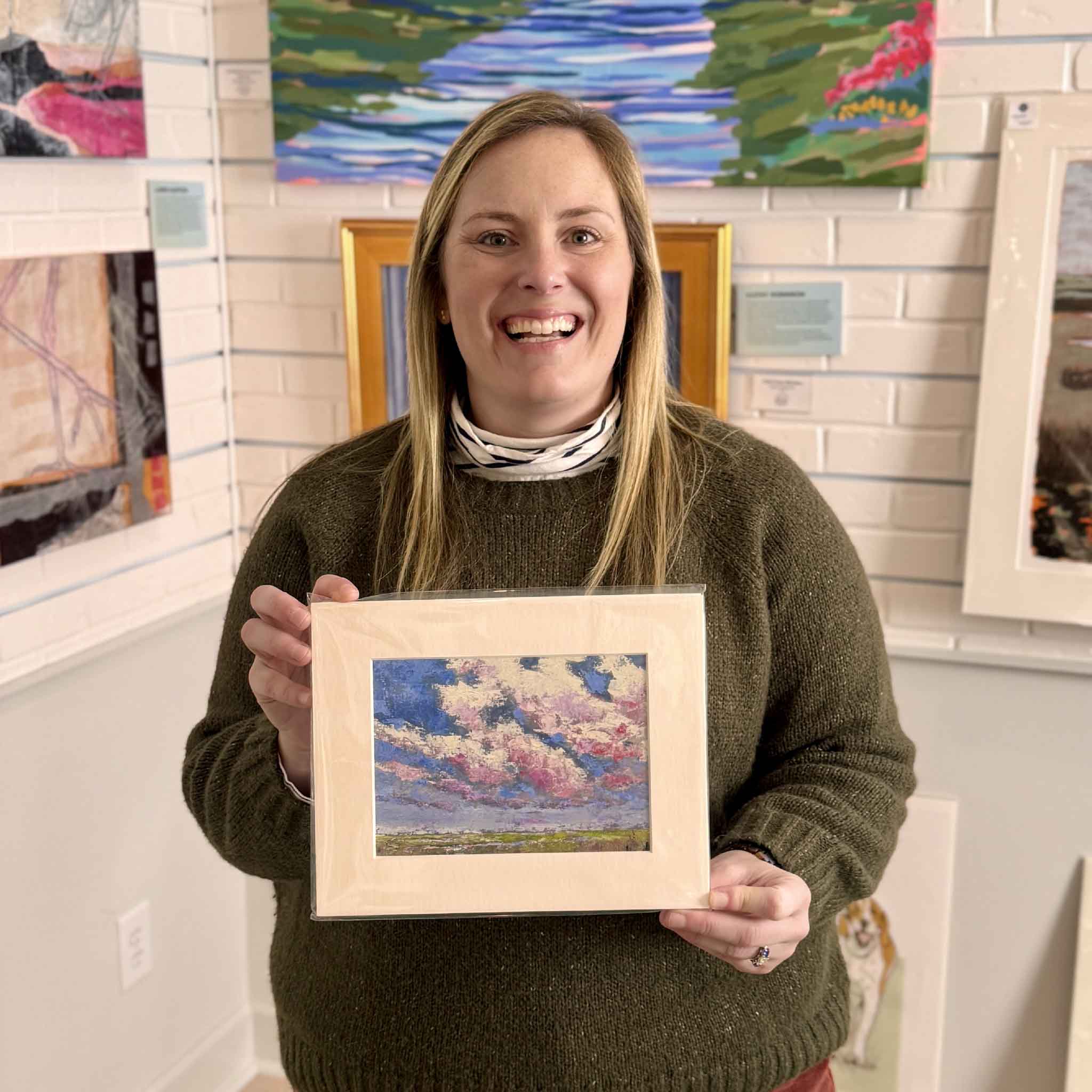 Gray Man Gallery team member Bordy holding the matted fine art print “Beneath the Blushing Sky” by Jeanne O’Neal inside the Pawleys Island gallery, showing scale and display style