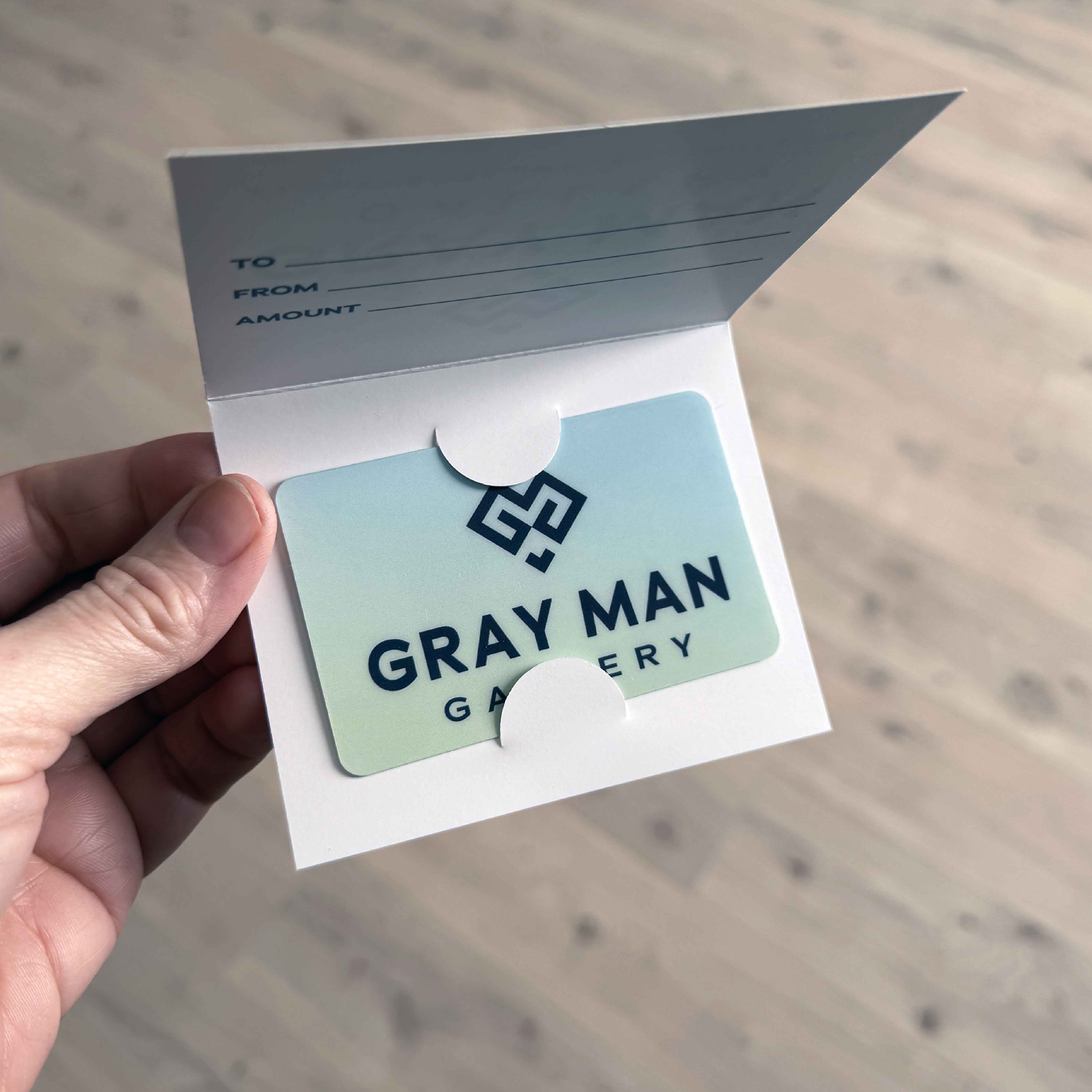 Gray Man Gallery gift card displayed inside a folded gift holder with To/From/Amount lines, ready for gifting.