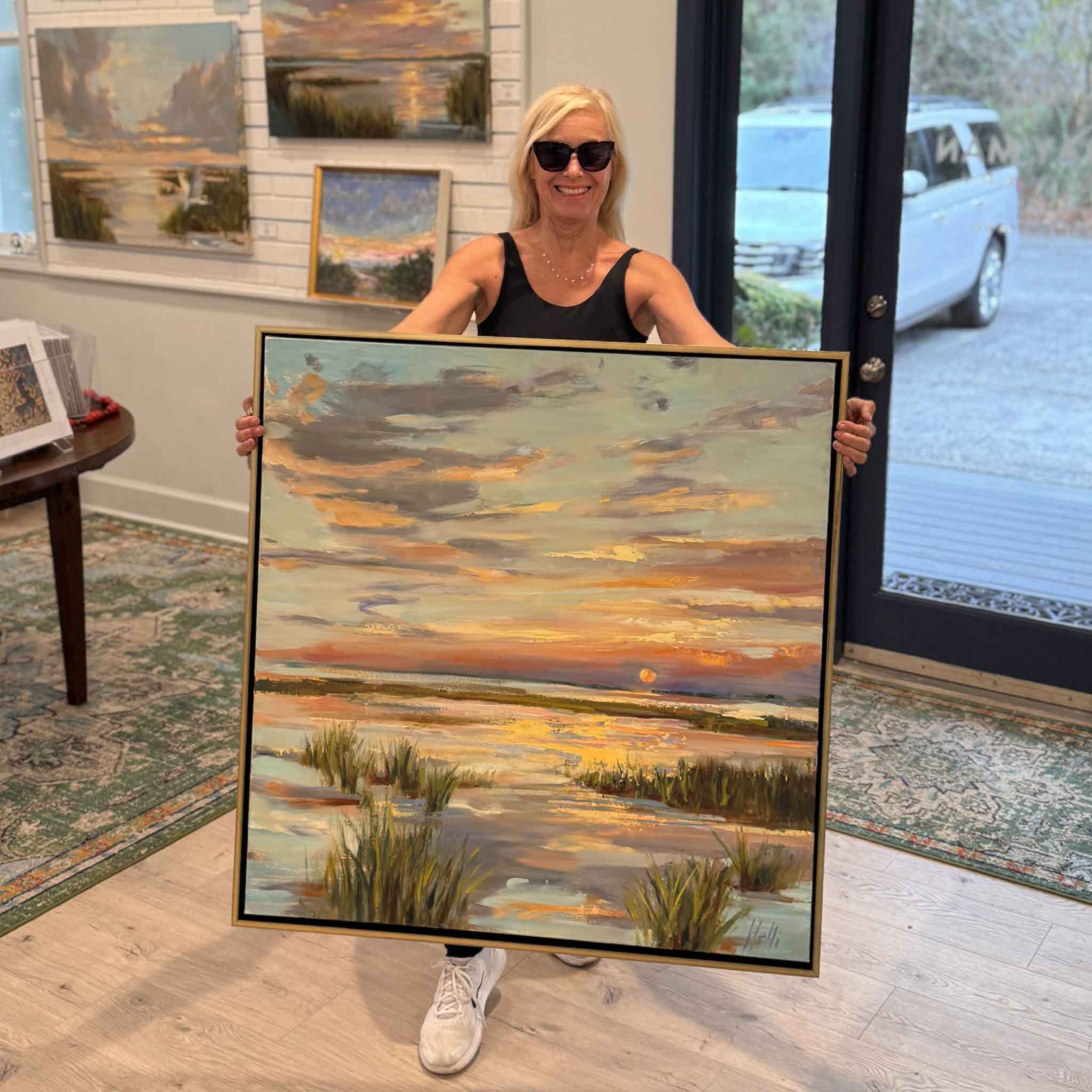 Lowcountry artist Helli Luck holding her original oil painting Evening Glow, a 36x36 marsh sunset with expressive brushwork and glowing horizon, inside Gray Man Gallery.