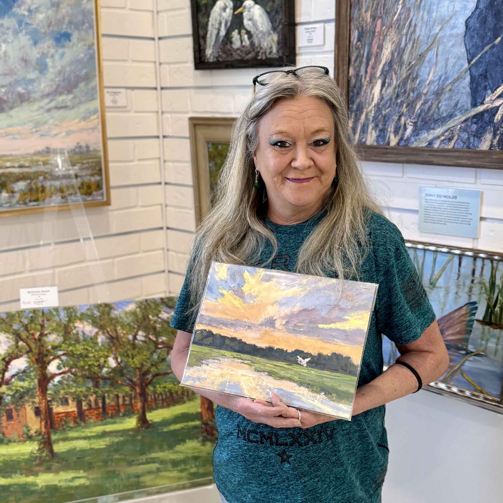 Charlotte holding Evening Grace by Jeanne O’Neal, a one-of-a-kind mixed-media artwork hand-painted in oil over a giclée print, depicting a Lowcountry marsh sunset with a white egret, sized 12x9 and ready for a standard frame, available at Gray Man Gallery.
