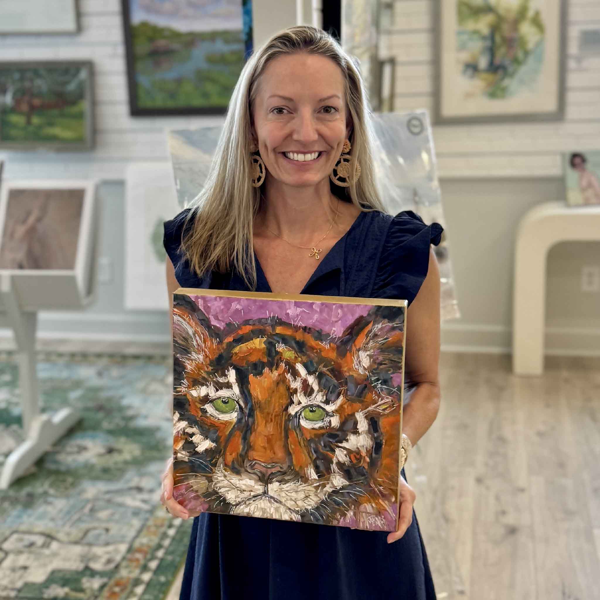 South Carolina artist Jeanne O'Neal holds her original oil painting Courage inside Gray Man Gallery in Pawleys Island, South Carolina. The expressive tiger portrait features vivid green eyes, bold orange and black brushwork, a vibrant violet background, and hand-painted gold gallery-wrapped edges, giving collectors a sense of the artwork's scale and the artist behind the piece.